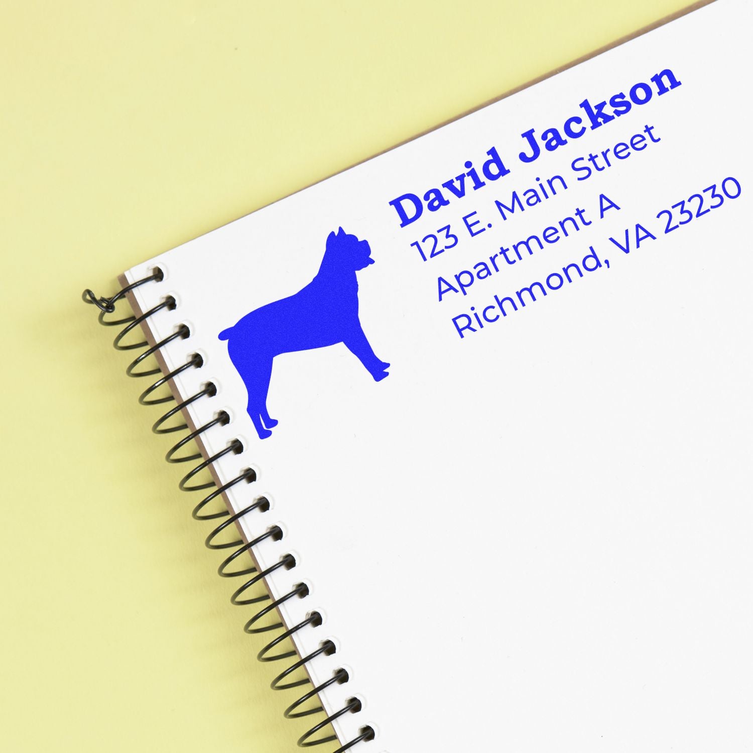 Cane Corso Silhouette Address Rubber Stamp on a notebook page, featuring a blue dog silhouette and sample address text in bold blue font on a light yellow background.