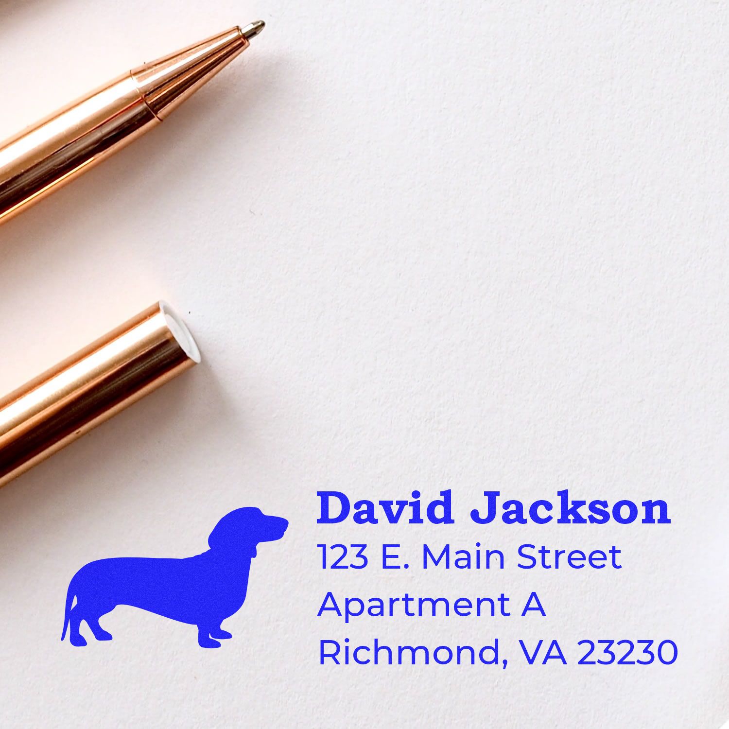 Self-Inking Dachshund Dog Address Stamp on paper with a blue dachshund silhouette, next to a gold pen. Text reads: David Jackson, 123 E. Main Street, Apartment A, Richmond, VA 23230.