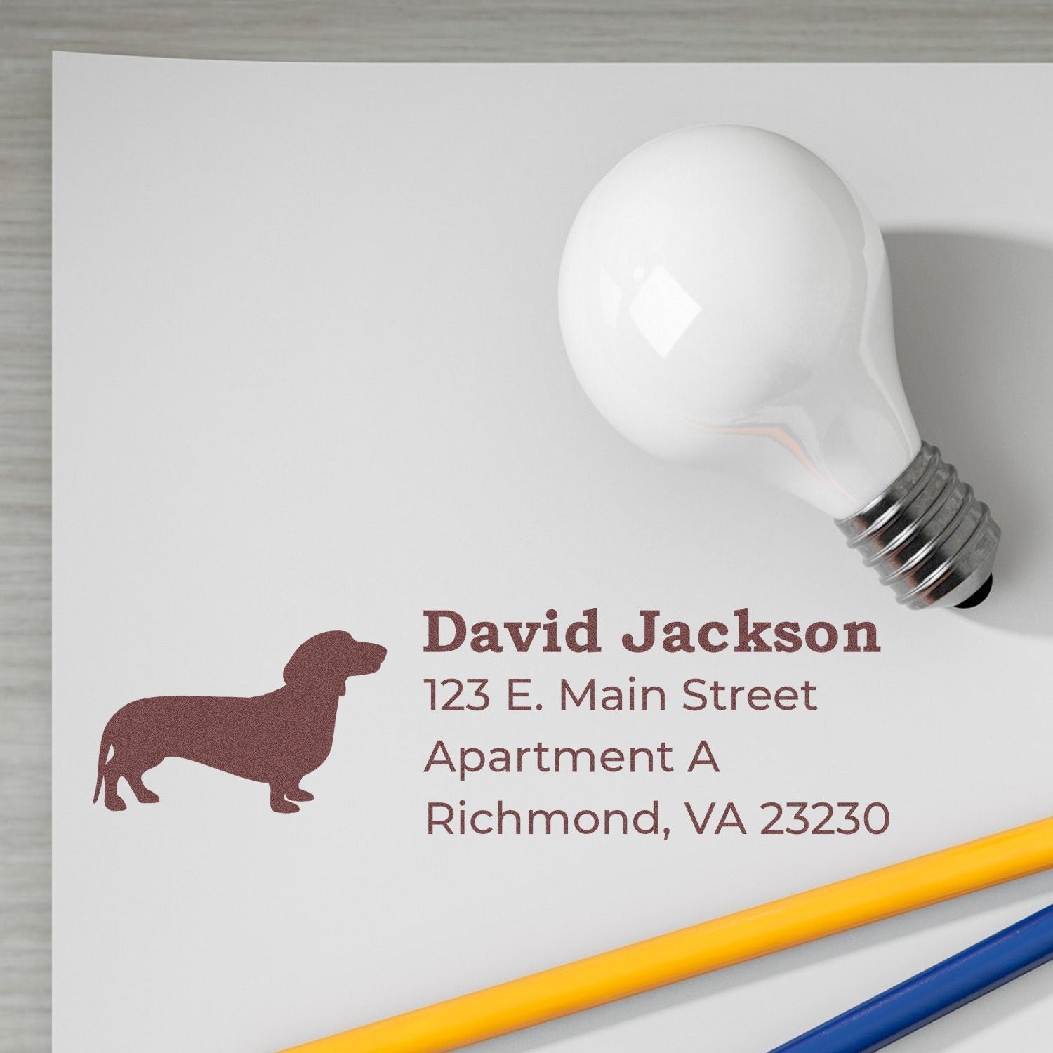 Slim Pre-Inked Dachshund Address Stamp on white paper with a dachshund silhouette, next to a light bulb and pencils. Text reads: David Jackson, 123 E. Main Street, Apartment A, Richmond, VA 23230.