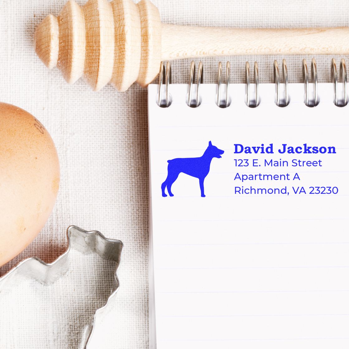 Self-Inking Doberman Dog Address Stamp on notepad with blue ink, next to a honey dipper, egg, and cookie cutter on a textured surface.