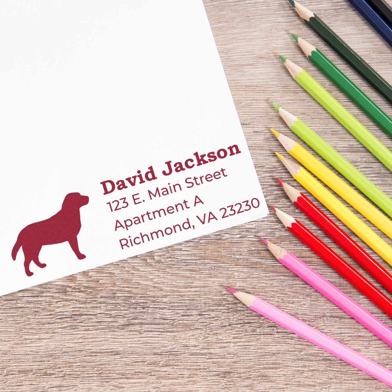 Labrador Silhouette Address Rubber Stamp on white paper with a red Labrador design, personalized address, and colorful pencils nearby on a wooden surface.