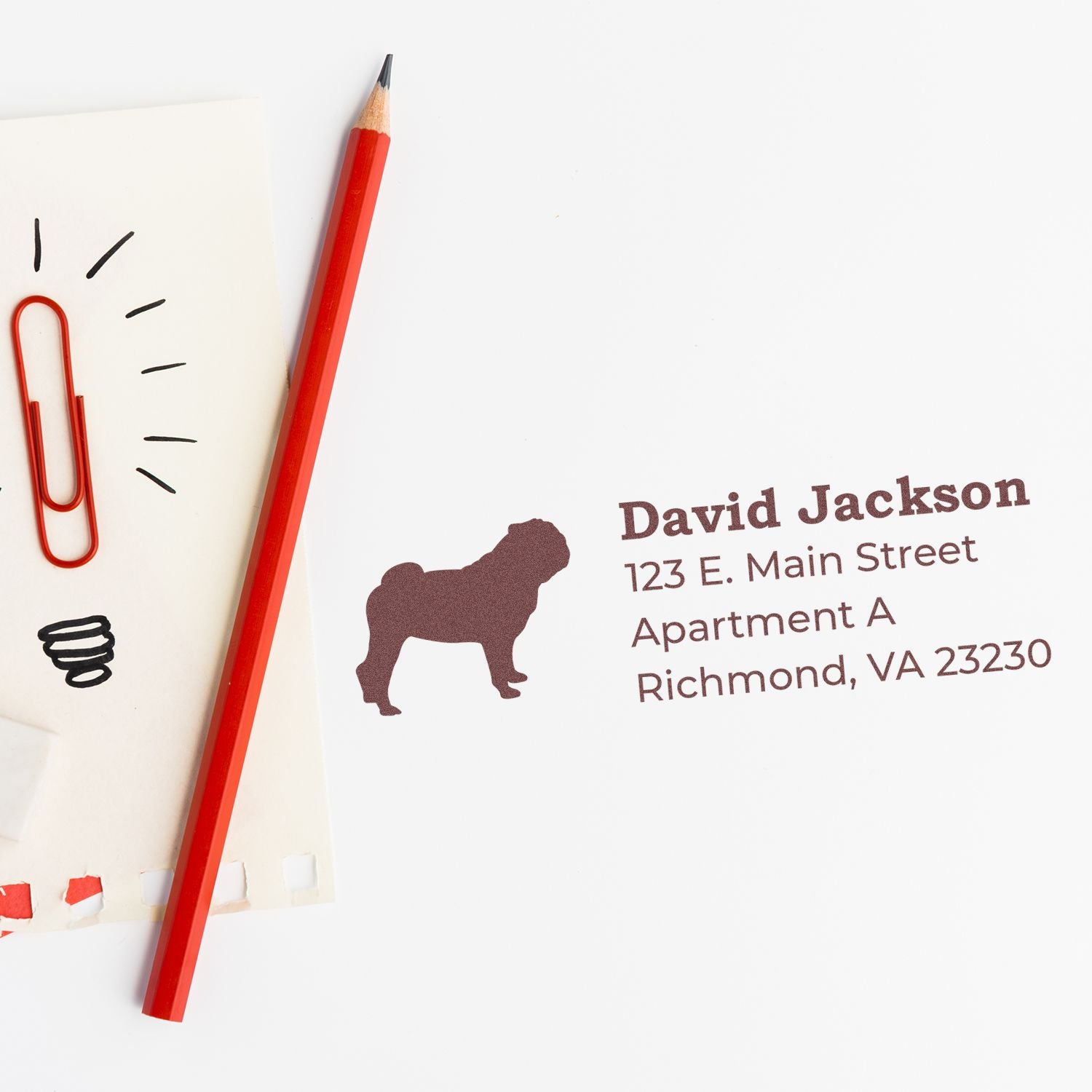 Self-Inking Pug Dog Address Stamp on white paper with a red pencil and paperclip. Displays a pug silhouette and sample address in brown ink. Perfect for personalized mail.