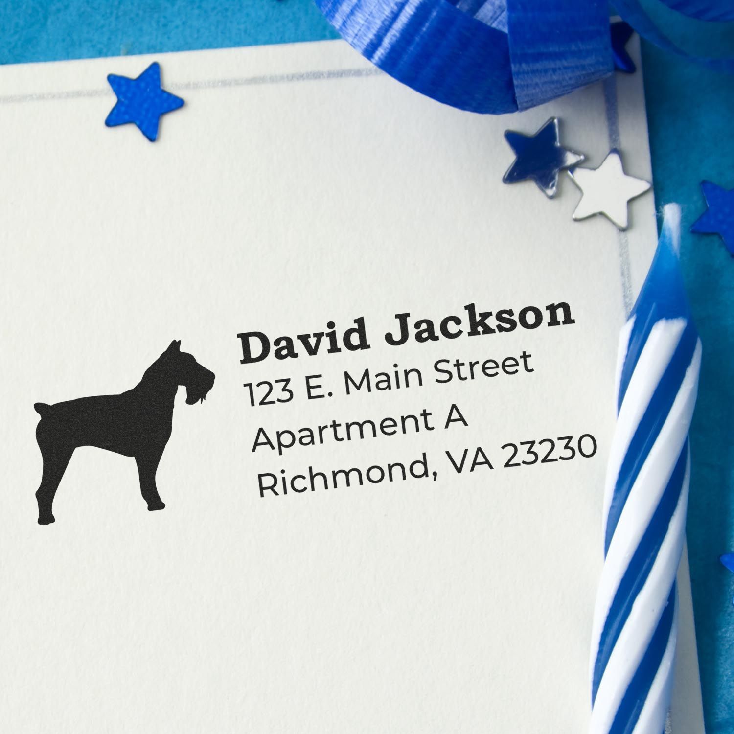 Slim Pre-Inked Schnauzer Address Stamp on an envelope with a schnauzer silhouette, surrounded by blue stars and ribbon, next to a blue and white striped candle.