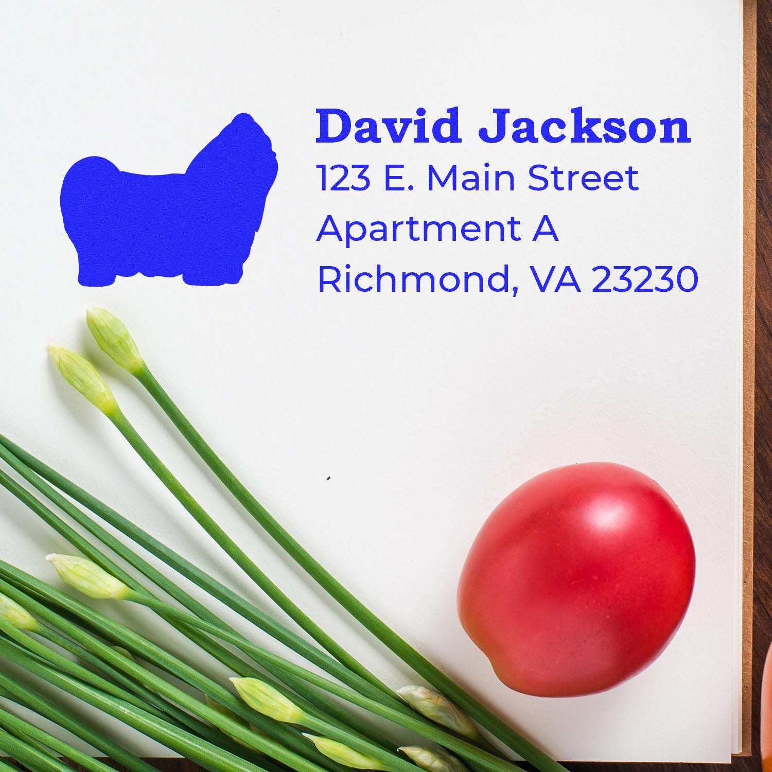 A PSI Pre-Inked Personalized Shih Tzu Address Stamp imprint on white paper, featuring a blue Shih Tzu silhouette and address text. Nearby are green onions and a red tomato on a wooden surface.