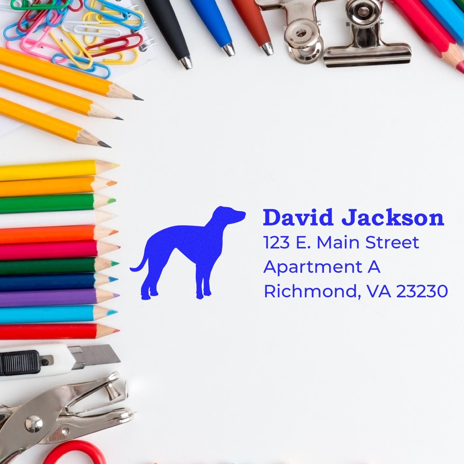 A Slim Pre-Inked Whippet Address Stamp on white paper, surrounded by colorful pencils, paperclips, and office supplies, displaying a blue whippet silhouette and sample address text.