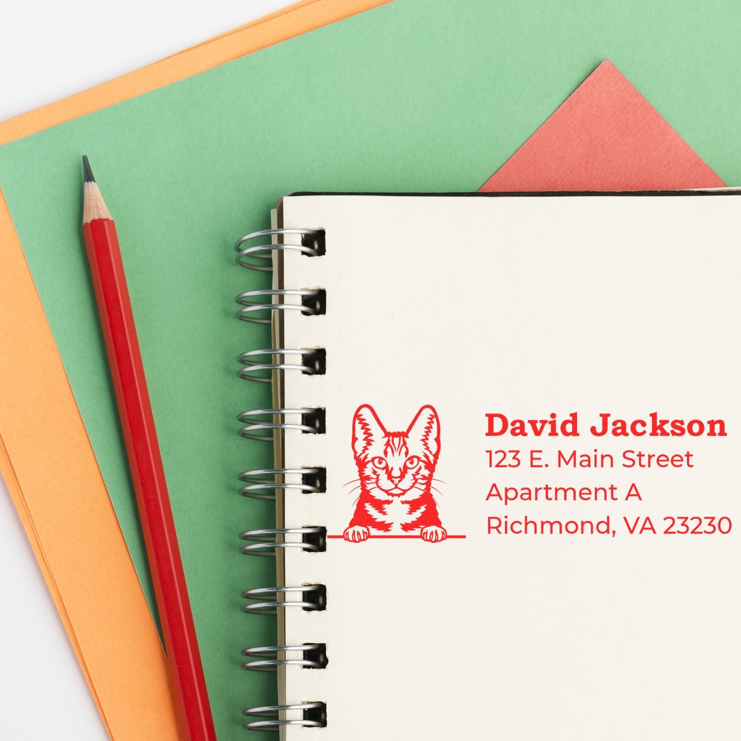 A notebook with a red Self-Inking American Shorthair Custom Address Stamp imprint, featuring a cat design, next to a red pencil on colorful paper.