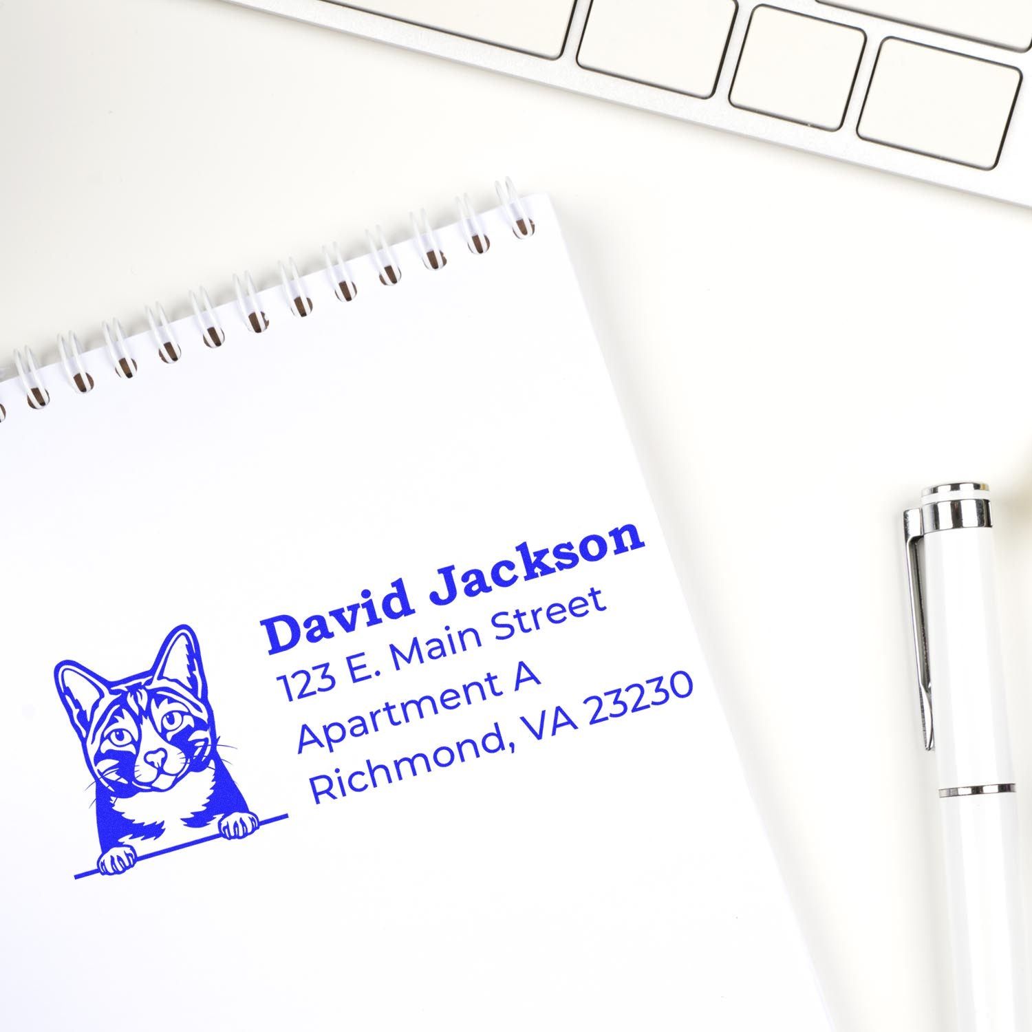 Slim Pre-Inked Arabian Mau Peeking Cat Return Address Stamp on a notepad next to a pen and keyboard, featuring a cute cat illustration and blue text for a personalized address.