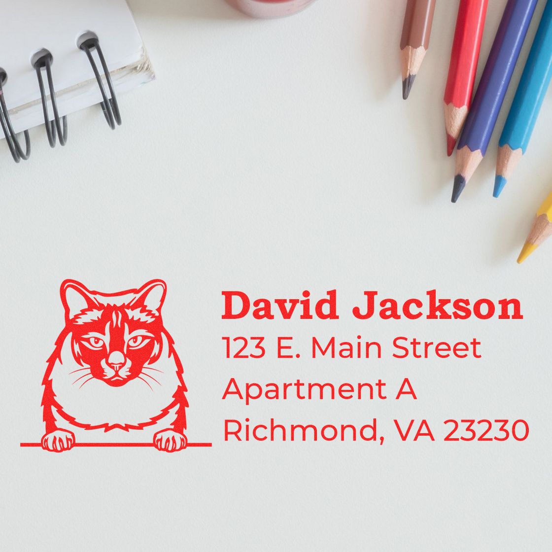 Wood Handle Balinese Cat Address Stamp on paper with a red cat design and address details. Nearby are colored pencils and a notebook, creating a creative and personalized stationery setting.