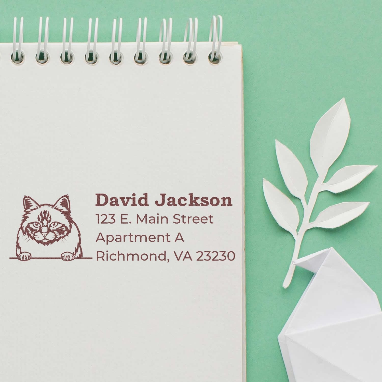 Self-Inking Birman Custom Address Stamp on a notepad with a cat design, displaying an address. Set against a green background with a paper leaf decoration.