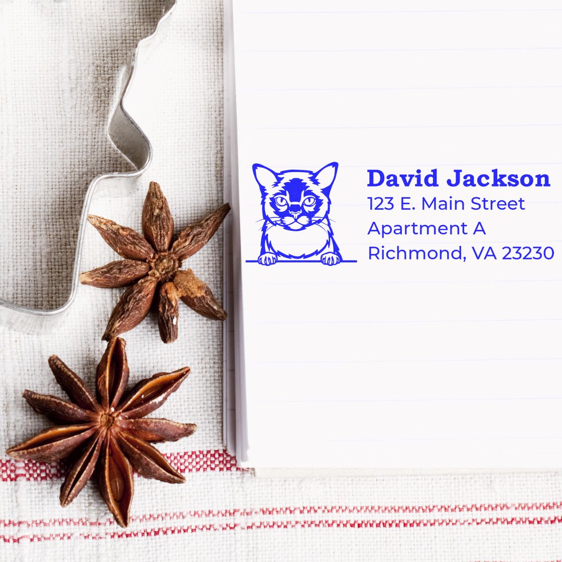 Wood Handle Burmese Cat Address Stamp on notepad with blue ink, featuring a cat illustration. Nearby are star anise and a cookie cutter on a striped cloth.