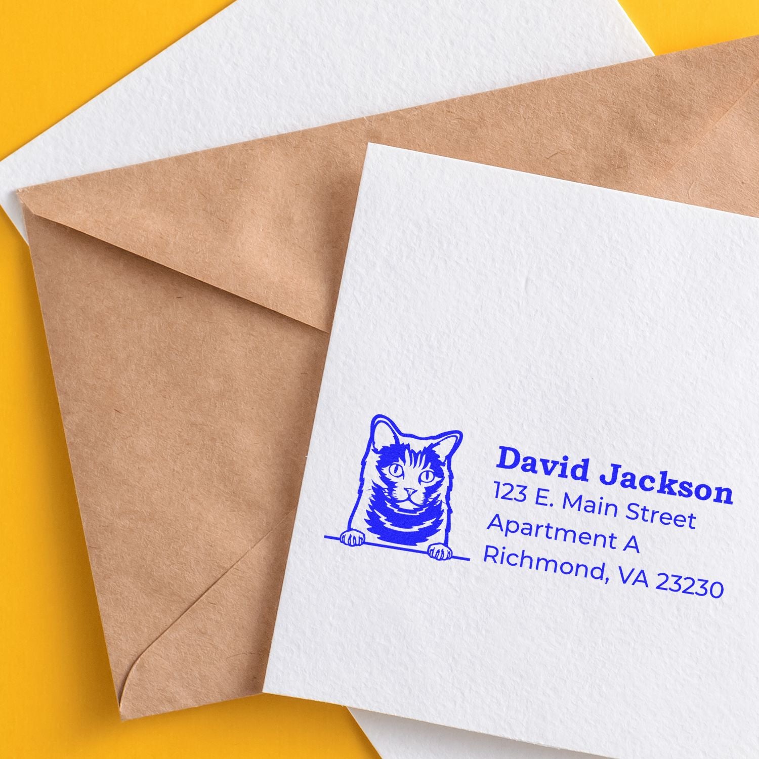 Wood Handle Chartreux Cat Address Stamp on white card with blue ink, featuring a cat illustration and sample address. Card rests on a brown envelope against a yellow background.