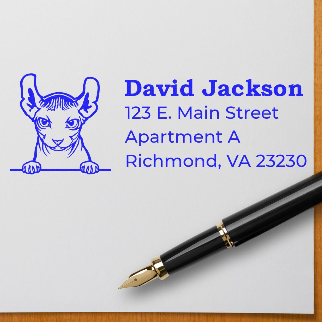 A Self-Inking Dwelf Custom Address Stamp on white paper with a blue cat design and sample address. A black fountain pen lies nearby on a wooden surface.