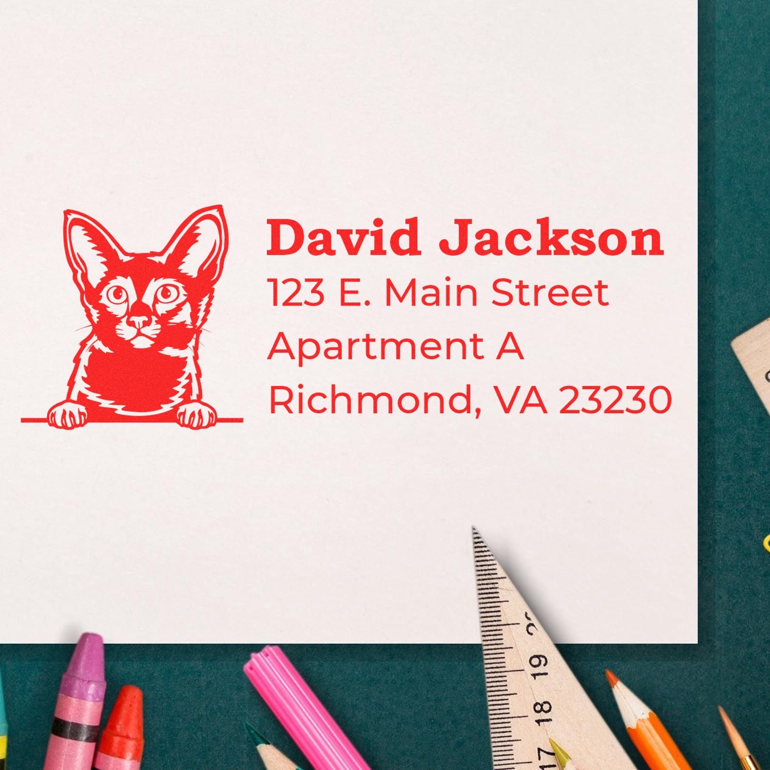 Self-Inking Javanese Custom Address Stamp on paper with a red cat design, displaying an address. Surrounded by colorful pencils and a ruler on a green surface.