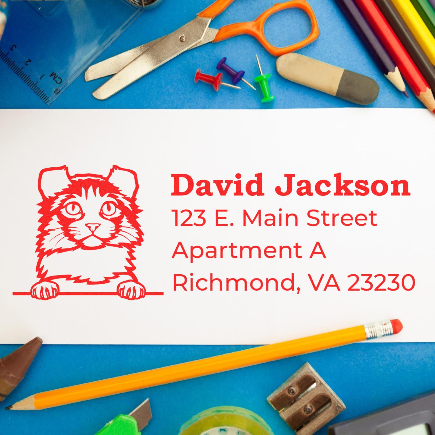 A Slim Pre-Inked Kinkalow Peeking Cat Return Address Stamp on an envelope, surrounded by colorful office supplies like scissors, push pins, and a ruler on a blue background.