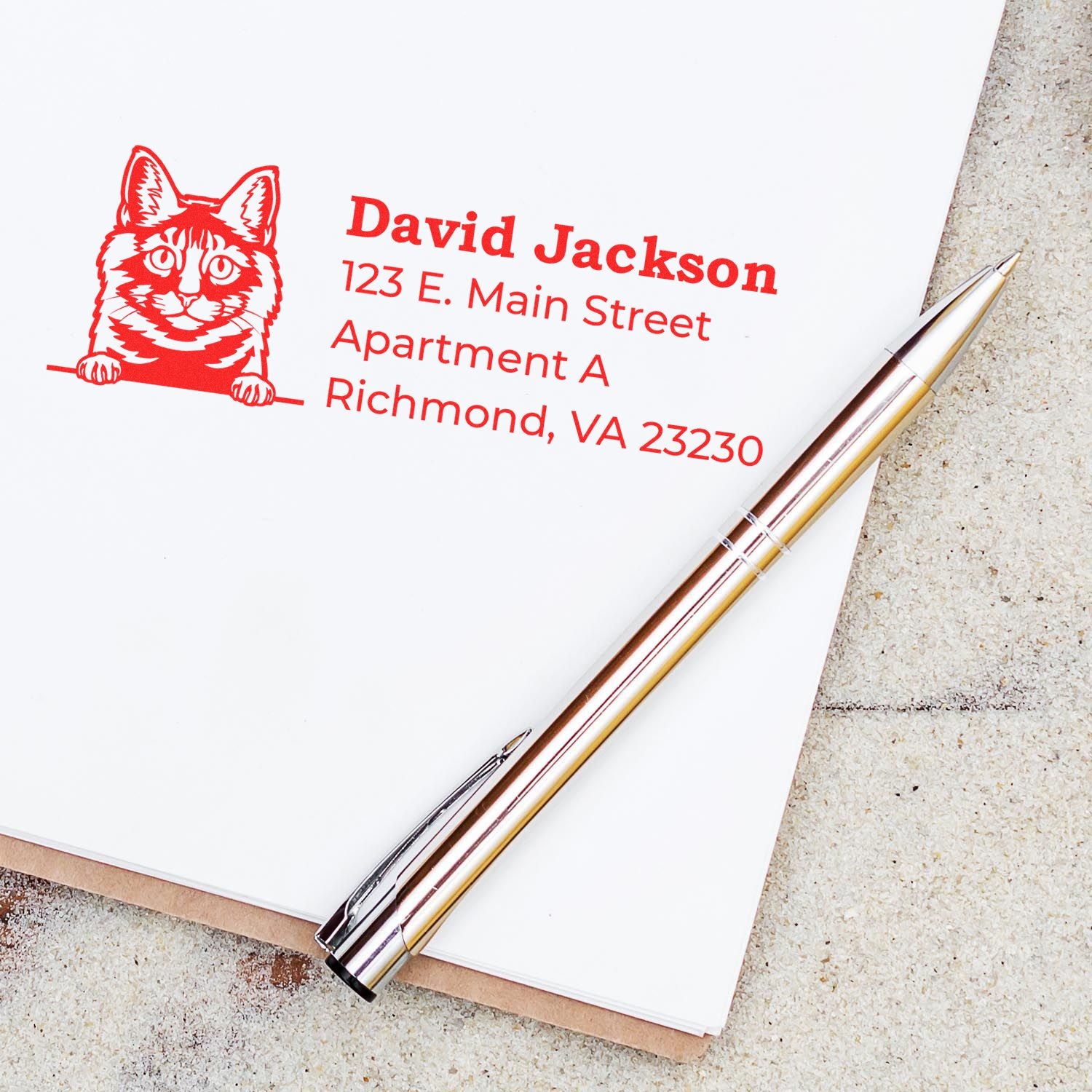 Slim Pre-Inked Kurilian Bobtail Peeking Cat Return Address Stamp in red ink on white paper, next to a silver pen. Features a cat illustration with address details.