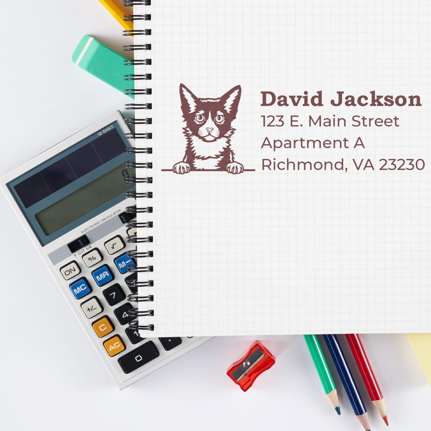 A Self-Inking LaPerm Custom Address Stamp on a notebook page with a cat design, next to a calculator, colored pencils, and a sharpener.