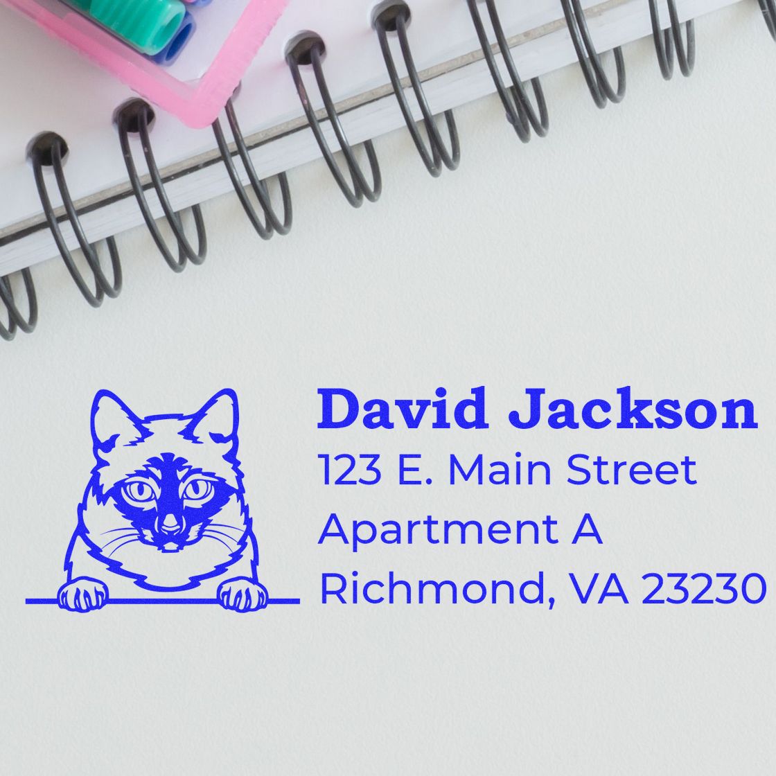 Slim Pre-Inked Mekong Bobtail Peeking Cat Return Address Stamp on paper with a blue cat design, next to a spiral notebook and colorful pens. Address reads: David Jackson, 123 E. Main Street, Richmond, VA.