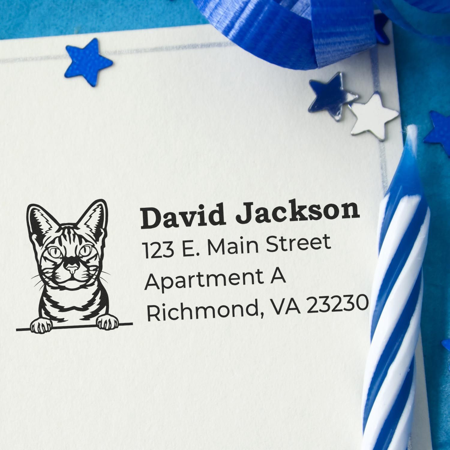 Wood Handle Ocicat Cat Address Stamp on an envelope with a cat illustration. Blue ribbon, stars, and a striped candle decorate the scene, creating a festive atmosphere.