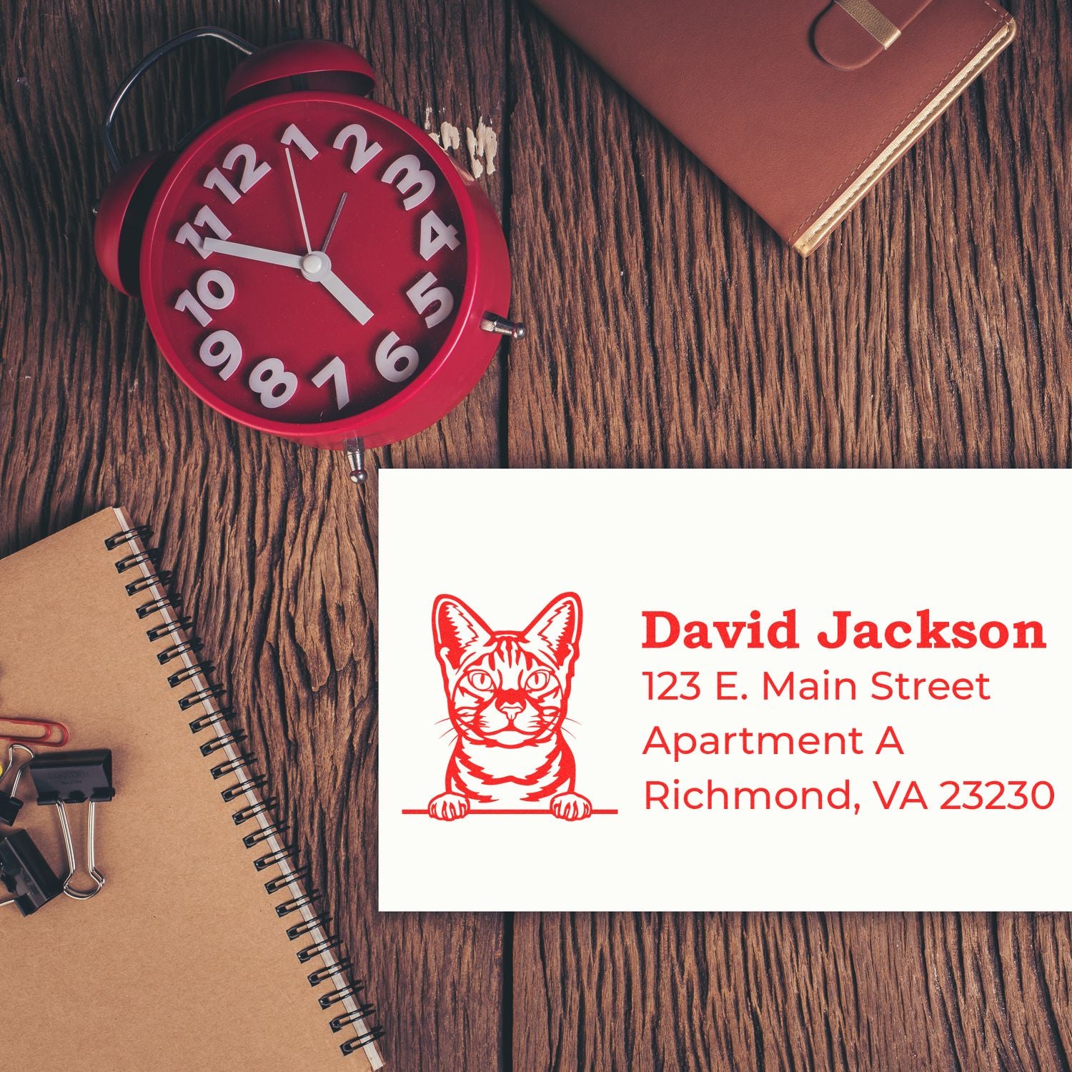 Wood Handle Ocicat Cat Address Stamp on a card with a red cat design, placed on a wooden desk with a red clock, notebook, and clips.