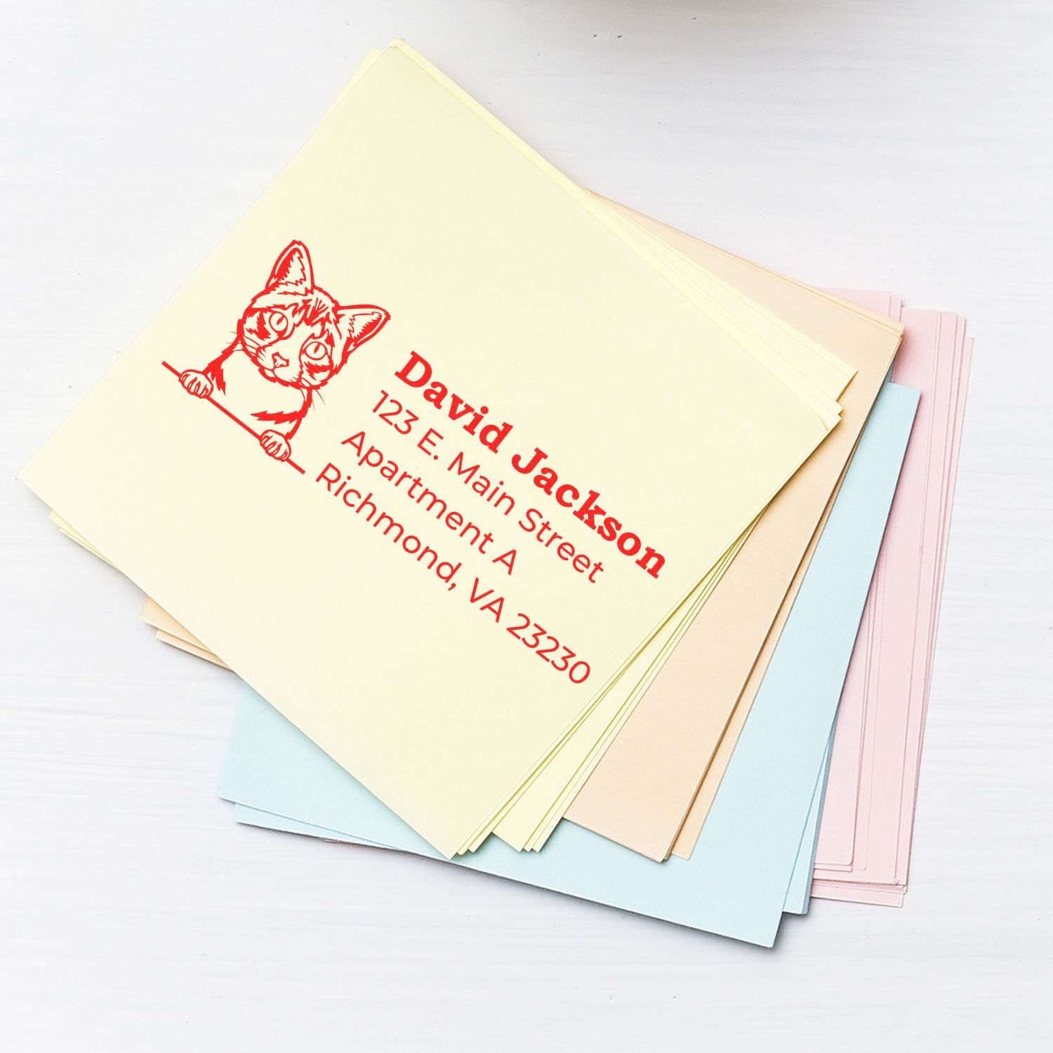 Stack of pastel envelopes with a yellow one on top, featuring a red cat design and personalized address from the PSI Pre-Inked Peeking Ojos Azules Cat Address Stamp.