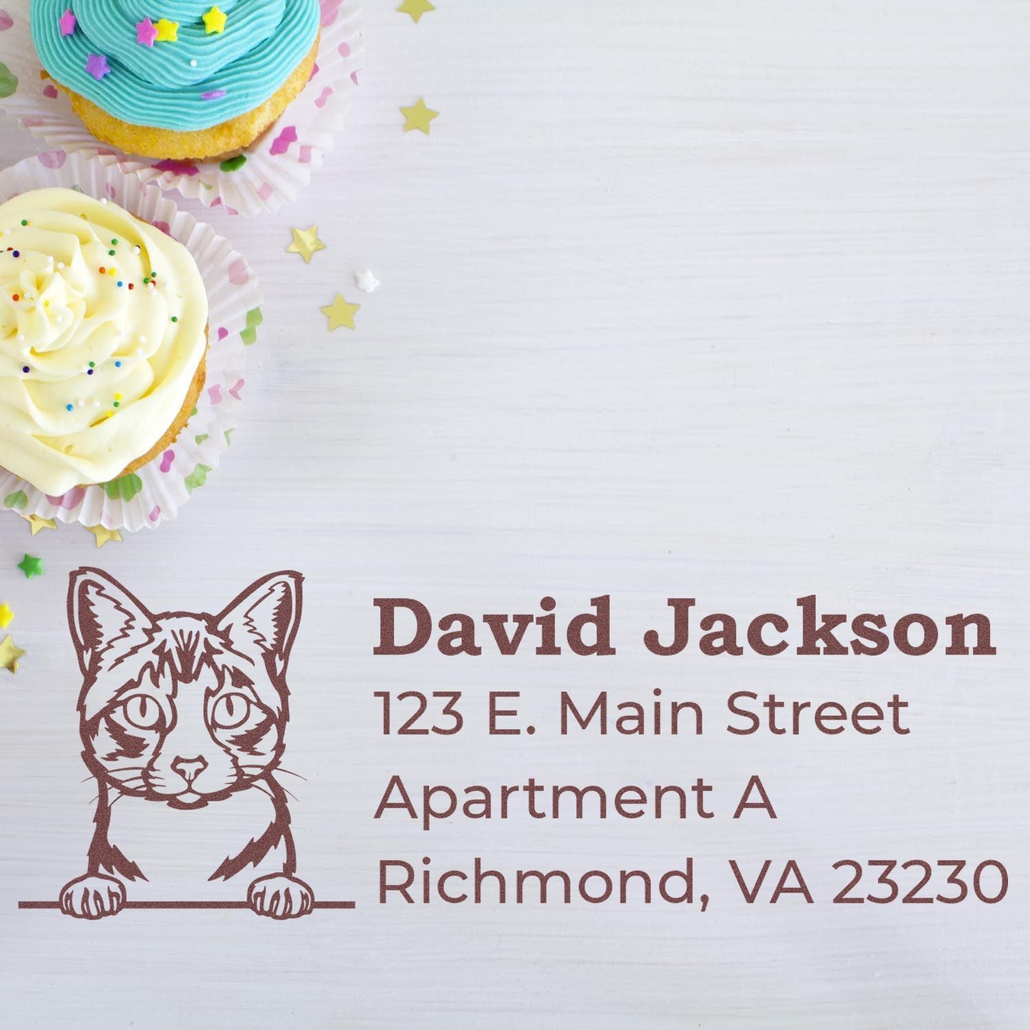 Cupcakes with sprinkles beside a stamped address featuring a cat illustration. Text reads David Jackson, 123 E. Main Street, Apartment A, Richmond, VA 23230. Self-Inking Ojos Azules Custom Address Stamp.