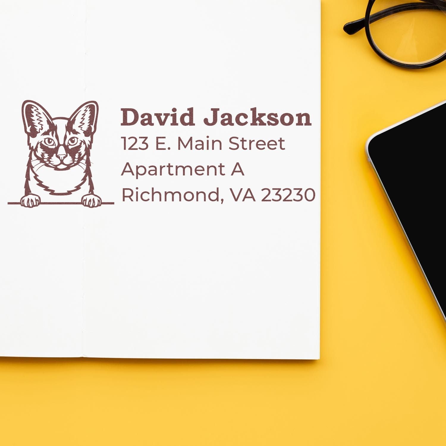 Self-Inking Oriental Longhair Custom Address Stamp on a white card with a cat illustration, next to glasses and a smartphone on a yellow surface.