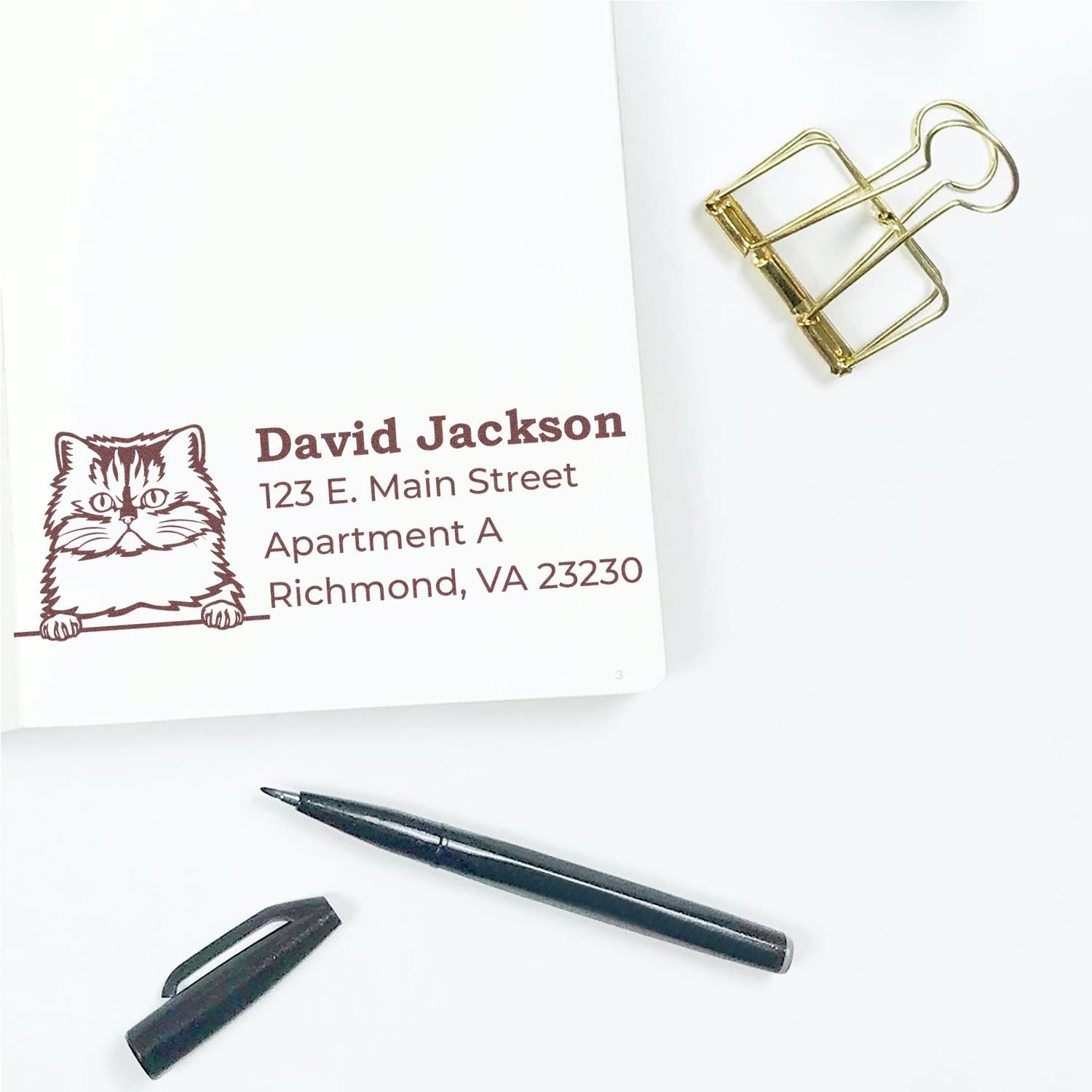 Wood Handle Persian Cat Address Stamp on white paper, featuring a cute cat illustration and sample address. Nearby are a black pen, pen cap, and gold binder clip on a white surface.