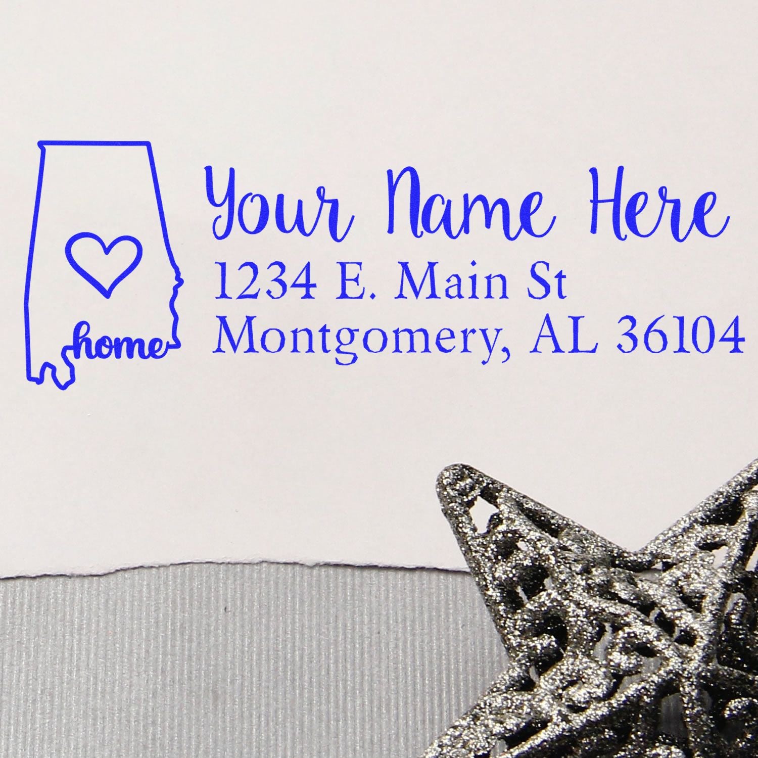 Alabama Self-Inking State Love Address Stamp on white paper with a blue outline of Alabama, heart symbol, and sample address. A decorative silver star is in the corner.