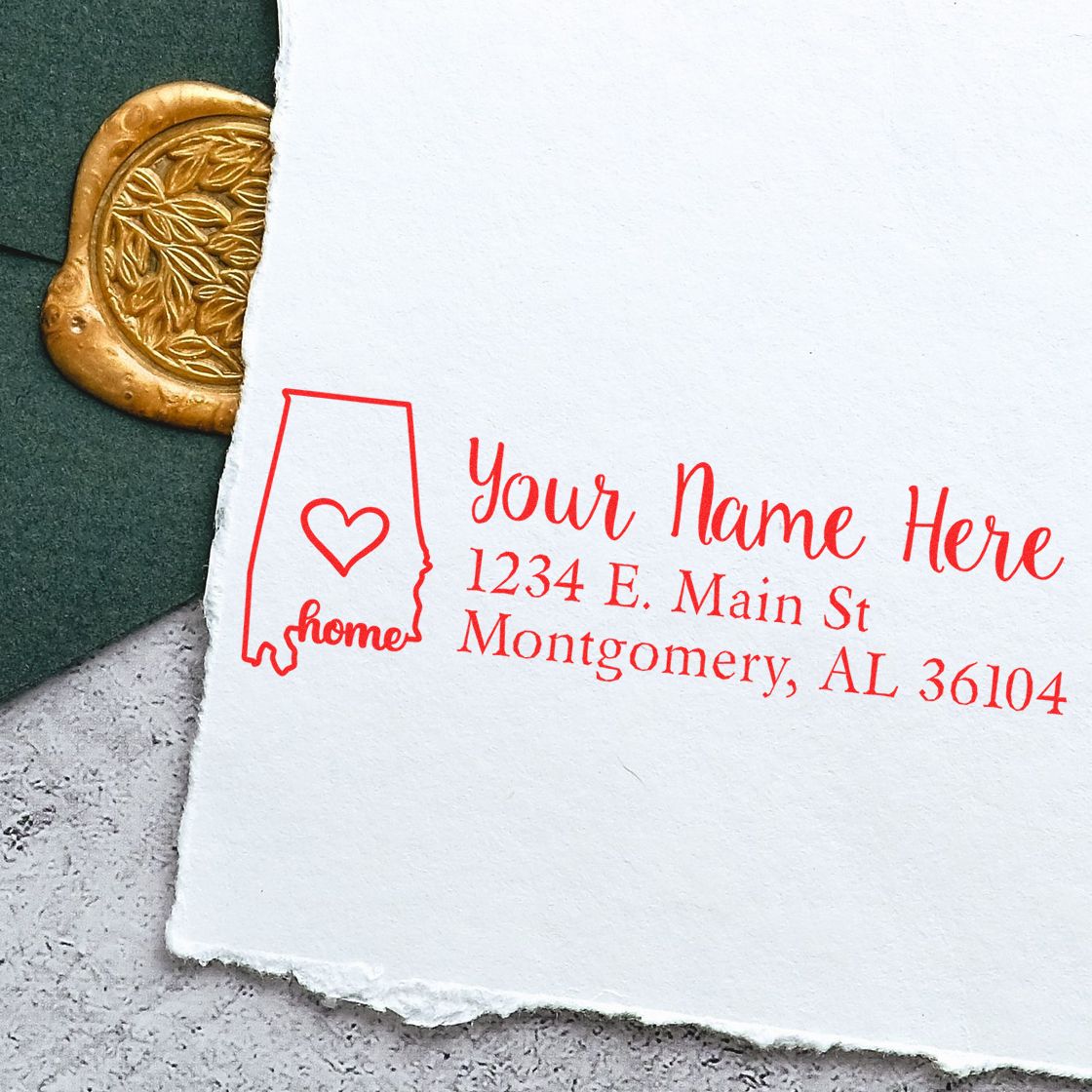 Wood Handle Alabama Custom Address Rubber Stamp imprint on white paper with a heart design, next to a green envelope with a gold wax seal. Text reads Your Name Here, 1234 E. Main St, Montgomery, AL 36104.