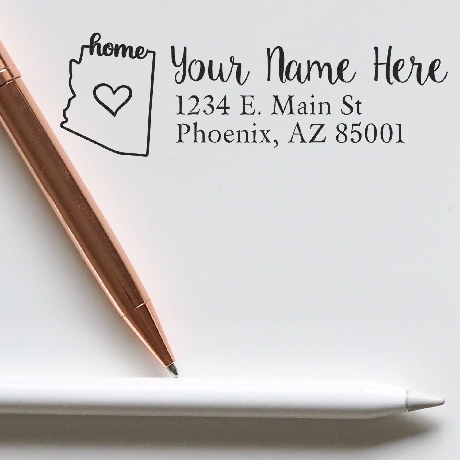 Slim Pre-Inked Arizona State Love Custom Address Stamp on white paper with a rose gold pen and a white pen, featuring a heart and home text inside the Arizona state outline.