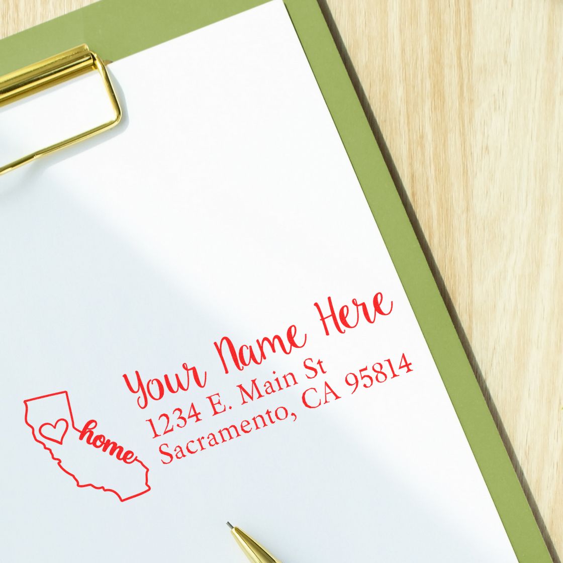 California Self-Inking State Love Address Stamp on paper with Your Name Here and a heart in the state outline. Includes address: 1234 E. Main St, Sacramento, CA 95814. Pen and clipboard visible.