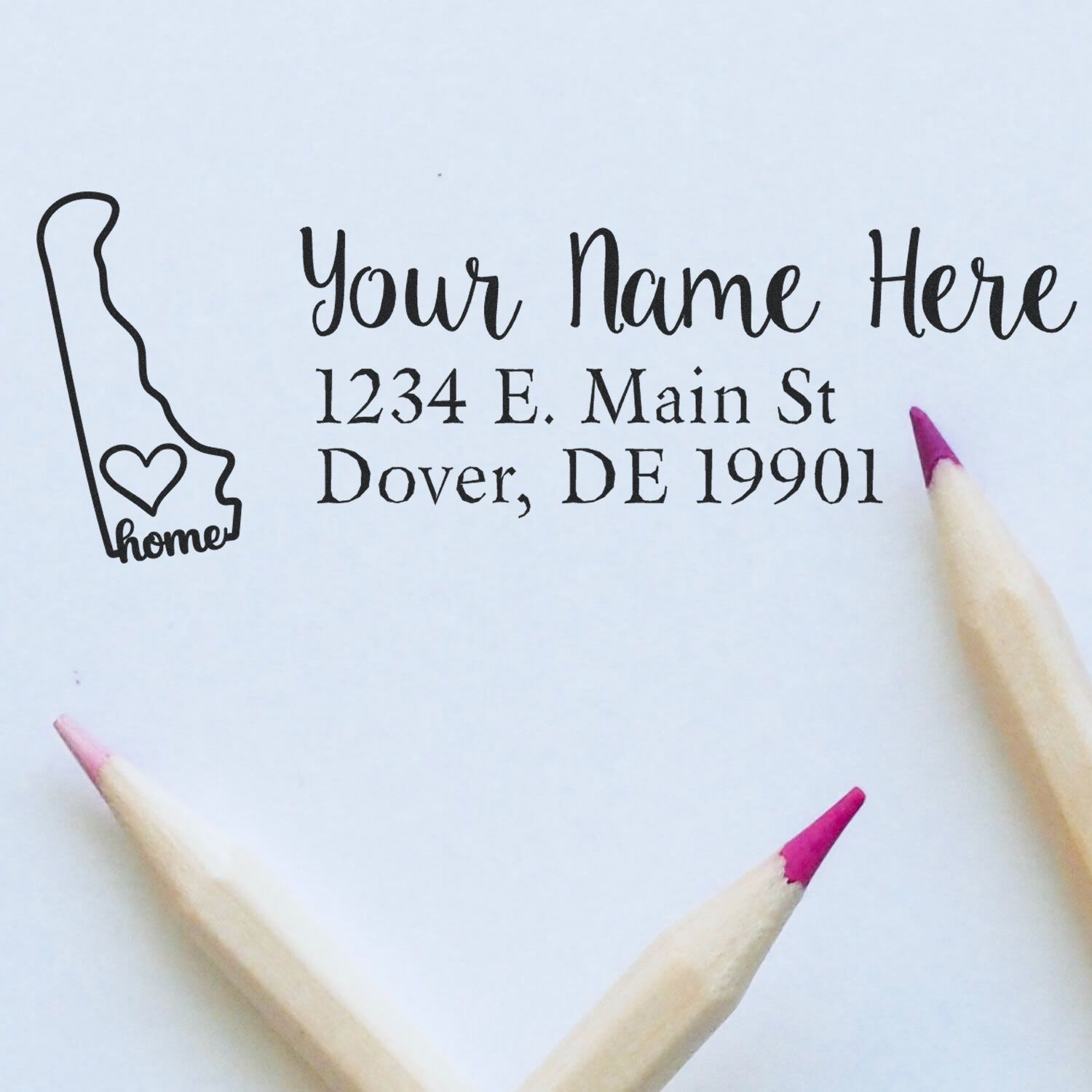 Delaware Self-Inking State Love Address Stamp on white paper with a heart design and sample address. Two pink-tipped pencils frame the image, highlighting the elegant, personalized stamp.