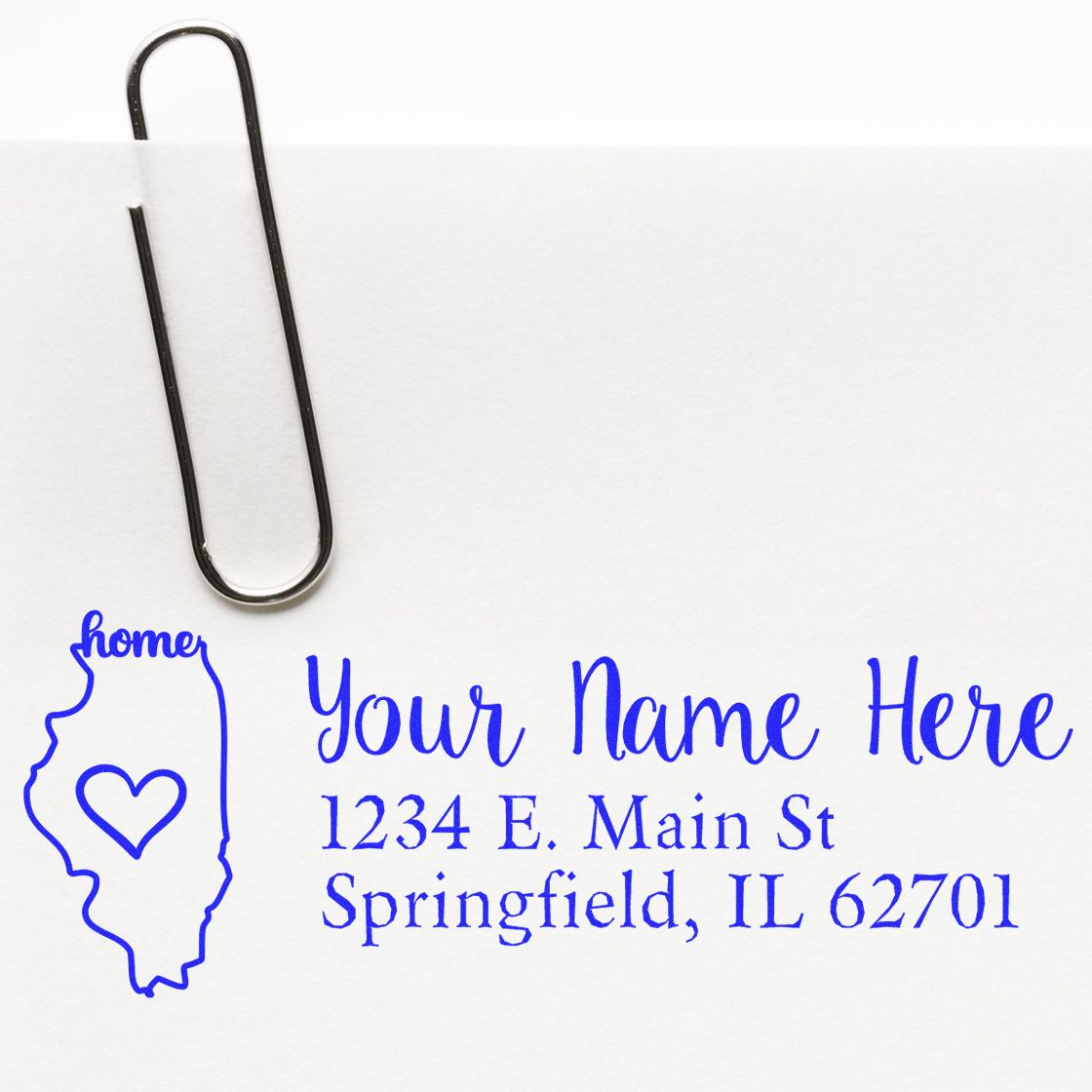 Wood Handle Illinois Custom Address Rubber Stamp on white paper with a blue ink design featuring a heart inside the state outline, next to a paperclip. Text reads Your Name Here, 1234 E. Main St, Springfield, IL 62701 .