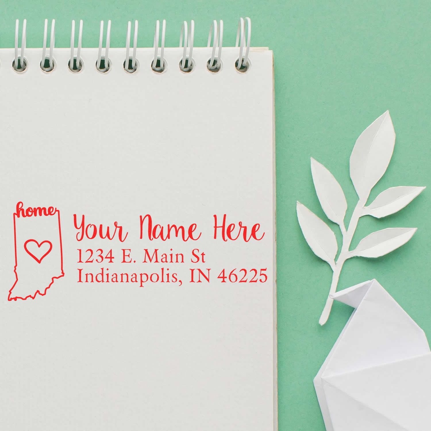 Indiana Self-Inking State Love Address Stamp on a notepad, featuring a red outline of Indiana with a heart and sample address text. Nearby are paper leaves and an envelope on a green background.