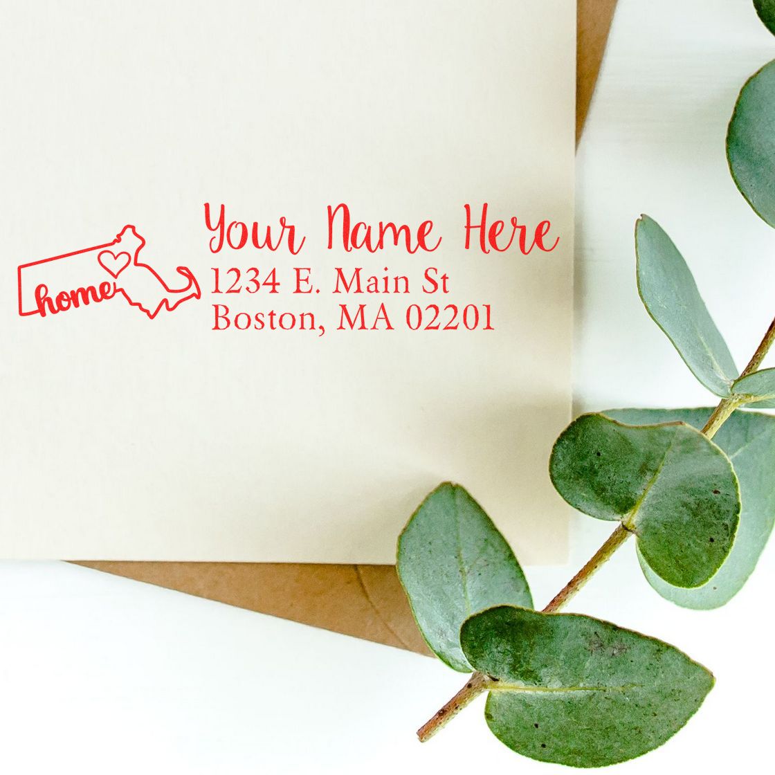 Envelope with red Massachusetts Self-Inking State Love Address Stamp, featuring a heart and state outline, next to eucalyptus leaves.