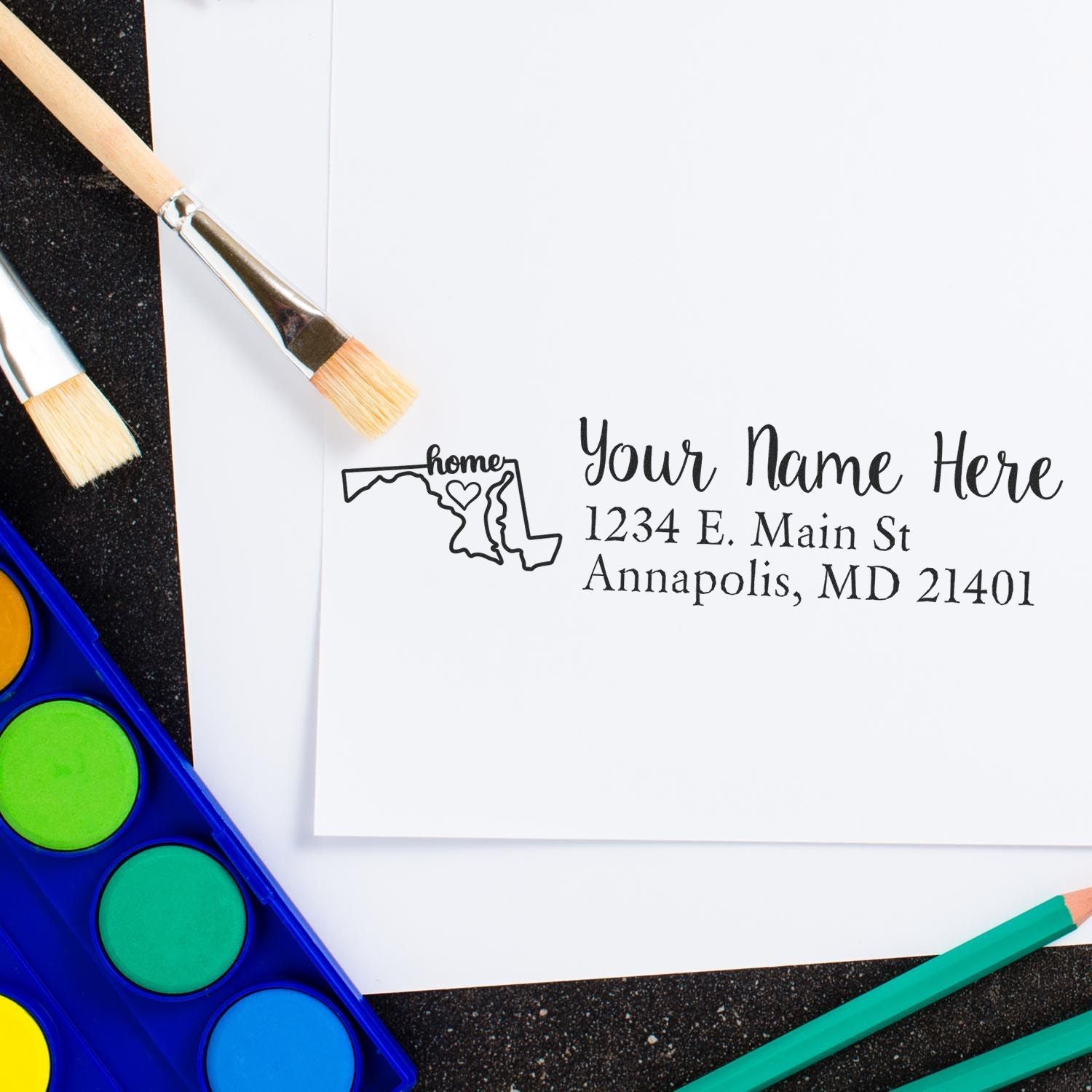 Maryland Self-Inking State Love Address Stamp on white paper with a Maryland outline and home text, surrounded by paintbrushes and watercolor paints.