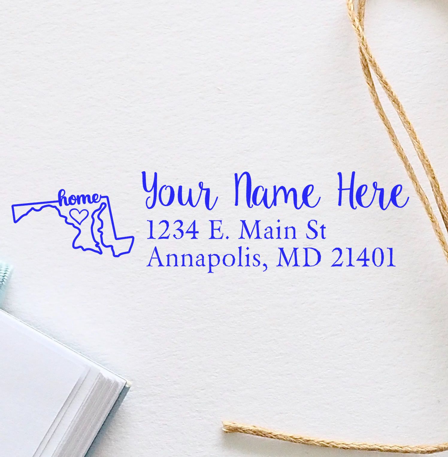 Wood Handle Maryland Custom Address Rubber Stamp on white paper, featuring a blue design with a map outline, heart, and sample address. Twine and a notebook are nearby.