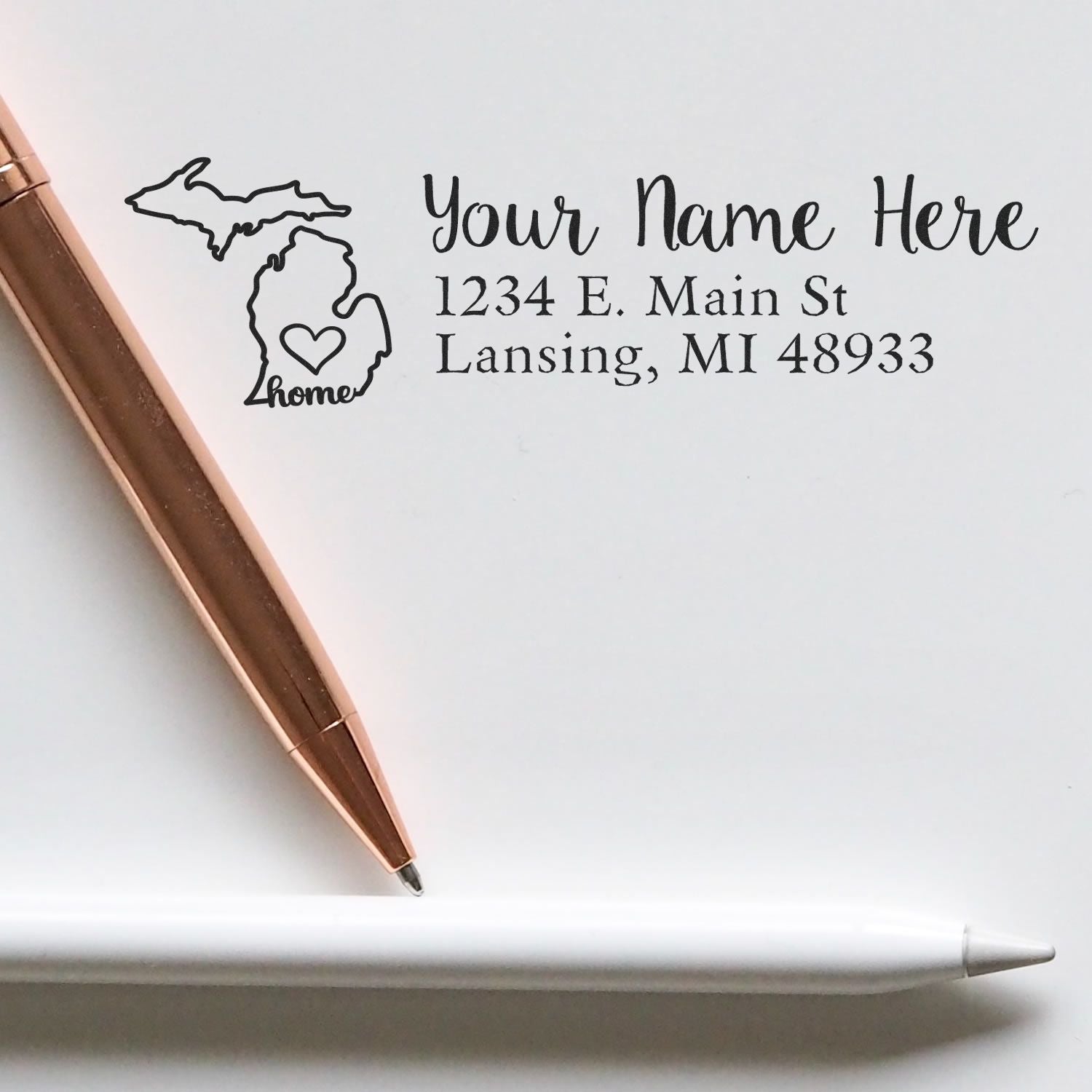 Michigan Self-Inking State Love Address Stamp on paper with a map outline and heart, next to a rose gold pen. Custom address: 1234 E. Main St, Lansing, MI 48933.