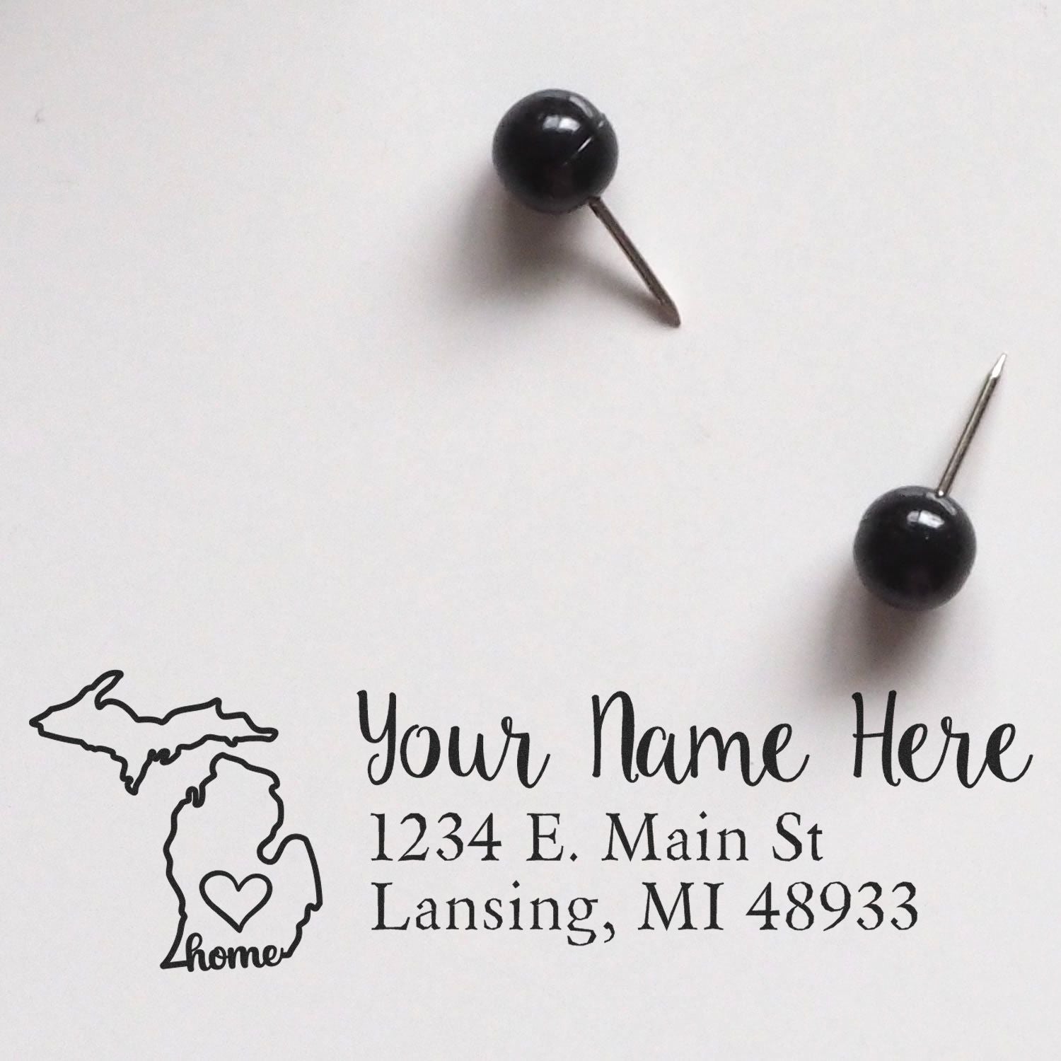 Michigan Self-Inking State Love Address Stamp on white paper with sample text Your Name Here and 1234 E. Main St, Lansing, MI 48933, next to two black push pins.