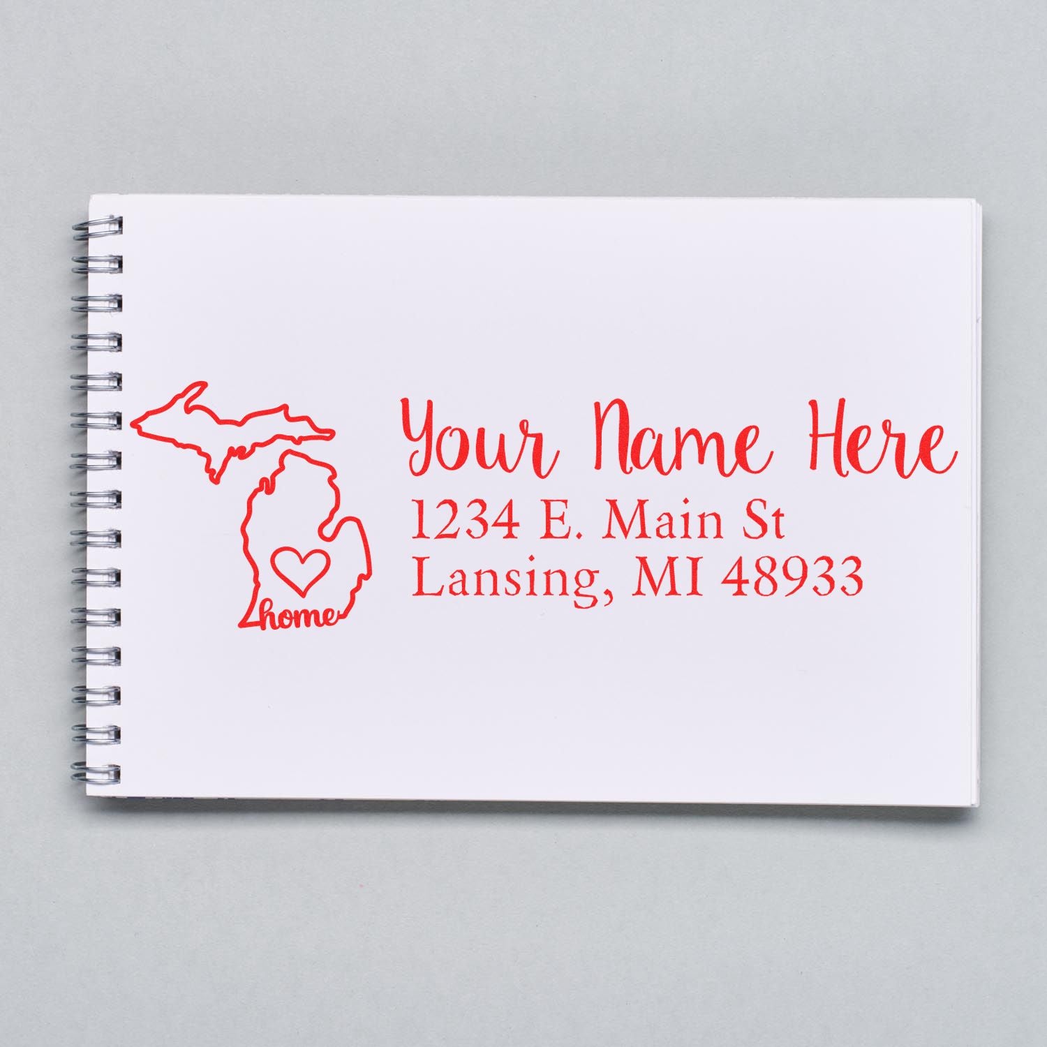 Notebook with a red Michigan Self-Inking State Love Address Stamp design, featuring a heart inside the state outline and customizable address text: Your Name Here, 1234 E. Main St, Lansing, MI 48933.