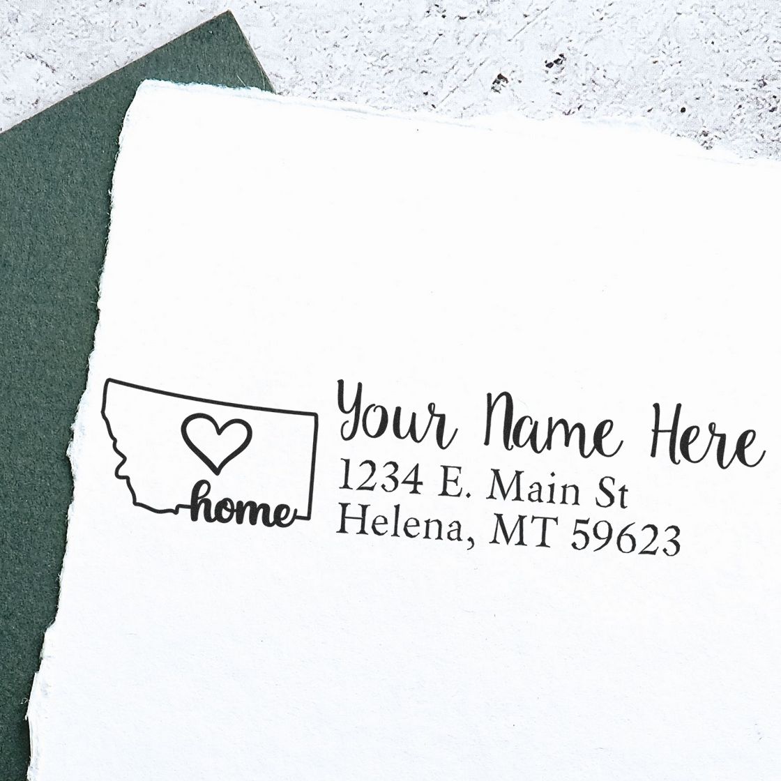 Montana Self-Inking State Love Address Stamp on white paper with a heart inside the state outline, personalized with Your Name Here, 1234 E. Main St, Helena, MT 59623, next to a green envelope.