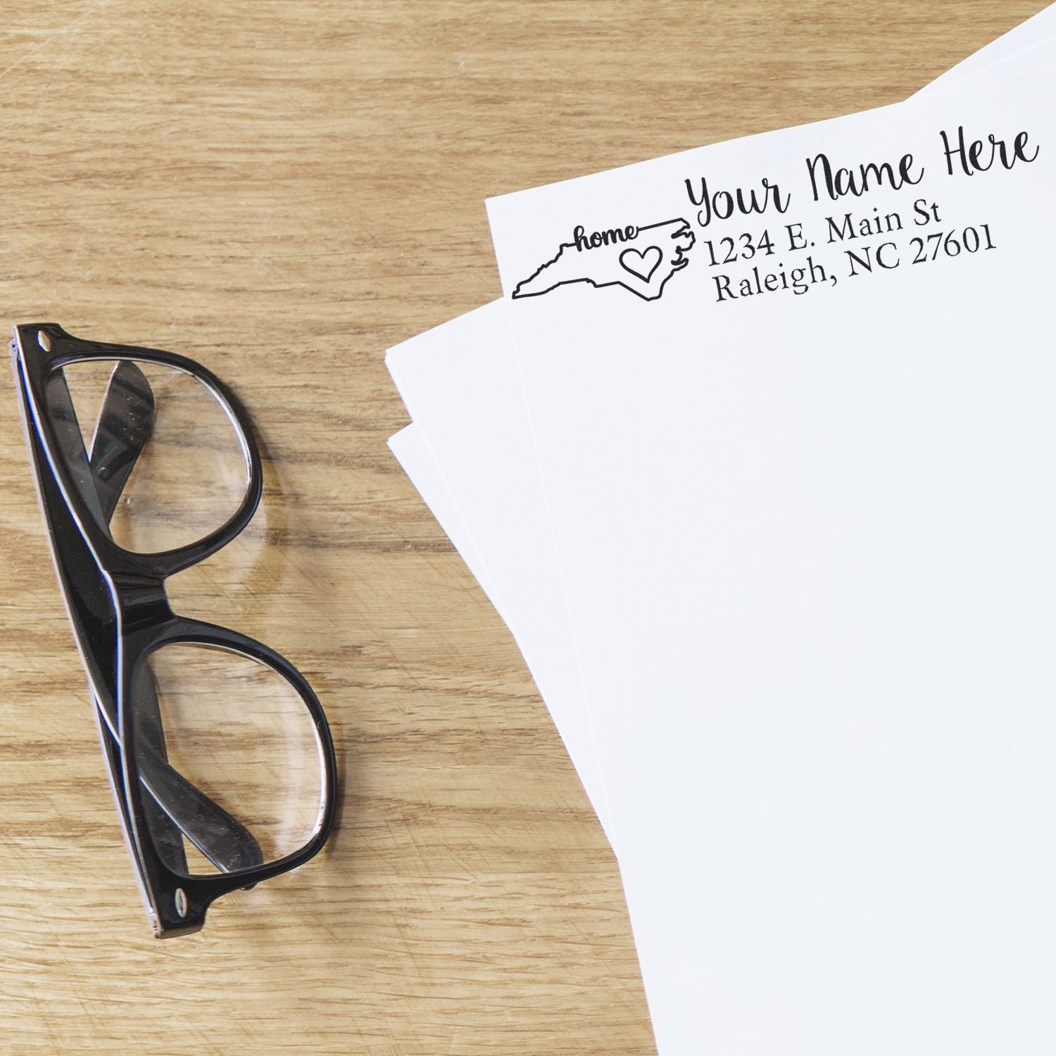 North Carolina Self-Inking State Love Address Stamp on white paper, featuring a heart design and sample address. Glasses rest nearby on a wooden surface.