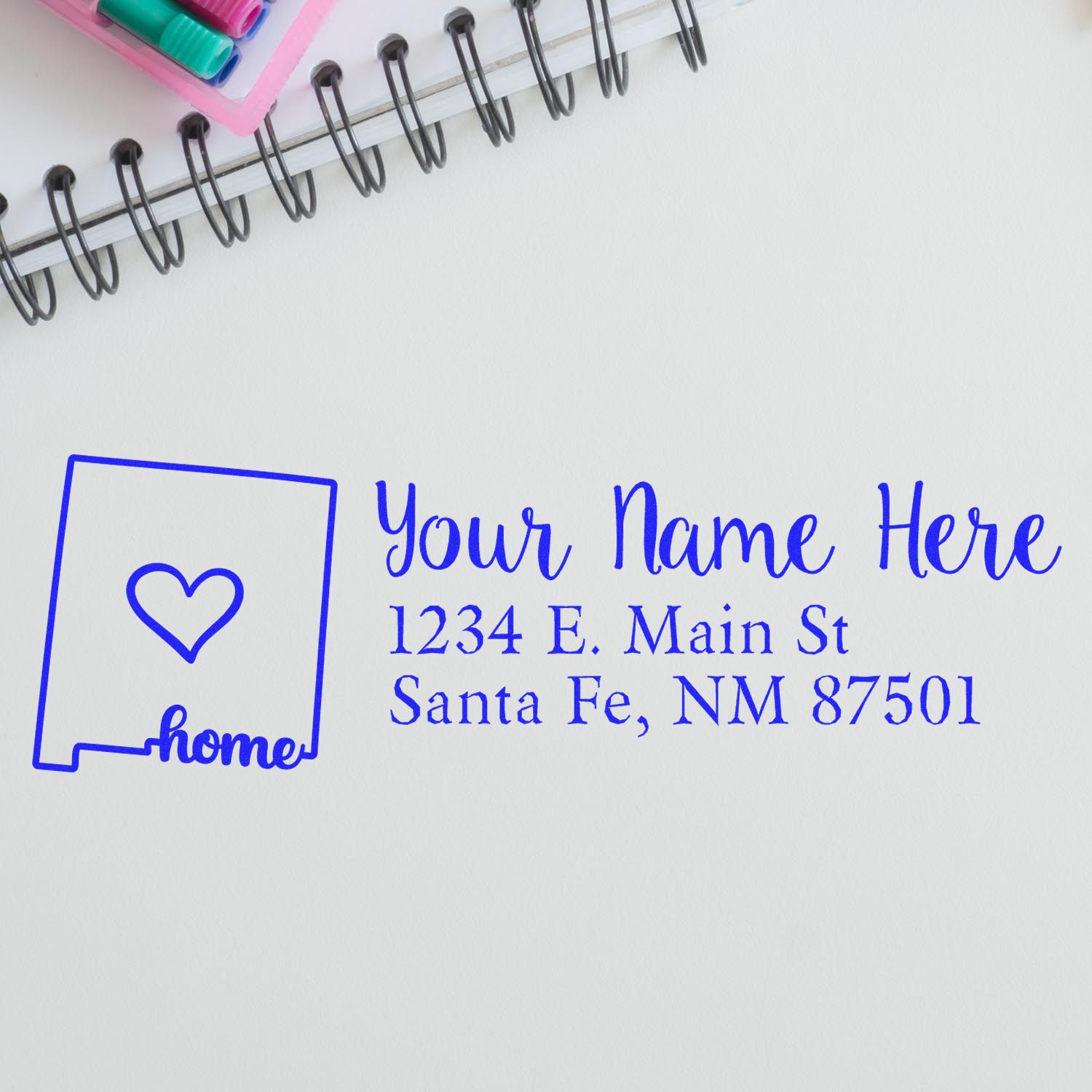 New Mexico Self-Inking State Love Address Stamp on paper with a heart design, personalized with name and address. Nearby are a notebook and colorful pens.