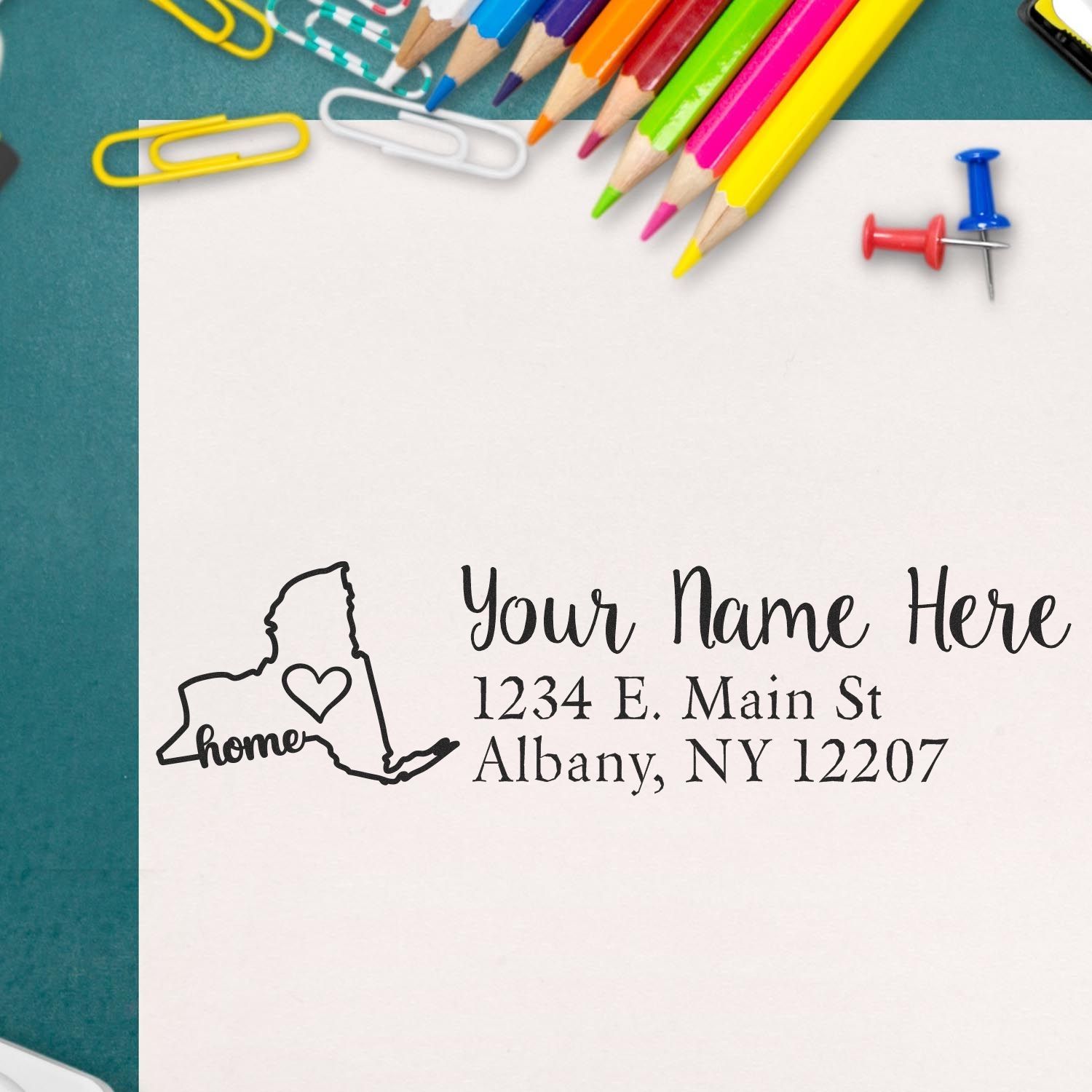 Slim Pre-Inked New York State Love Custom Address Stamp on a white envelope, surrounded by colorful pencils, paperclips, and pushpins on a green surface.