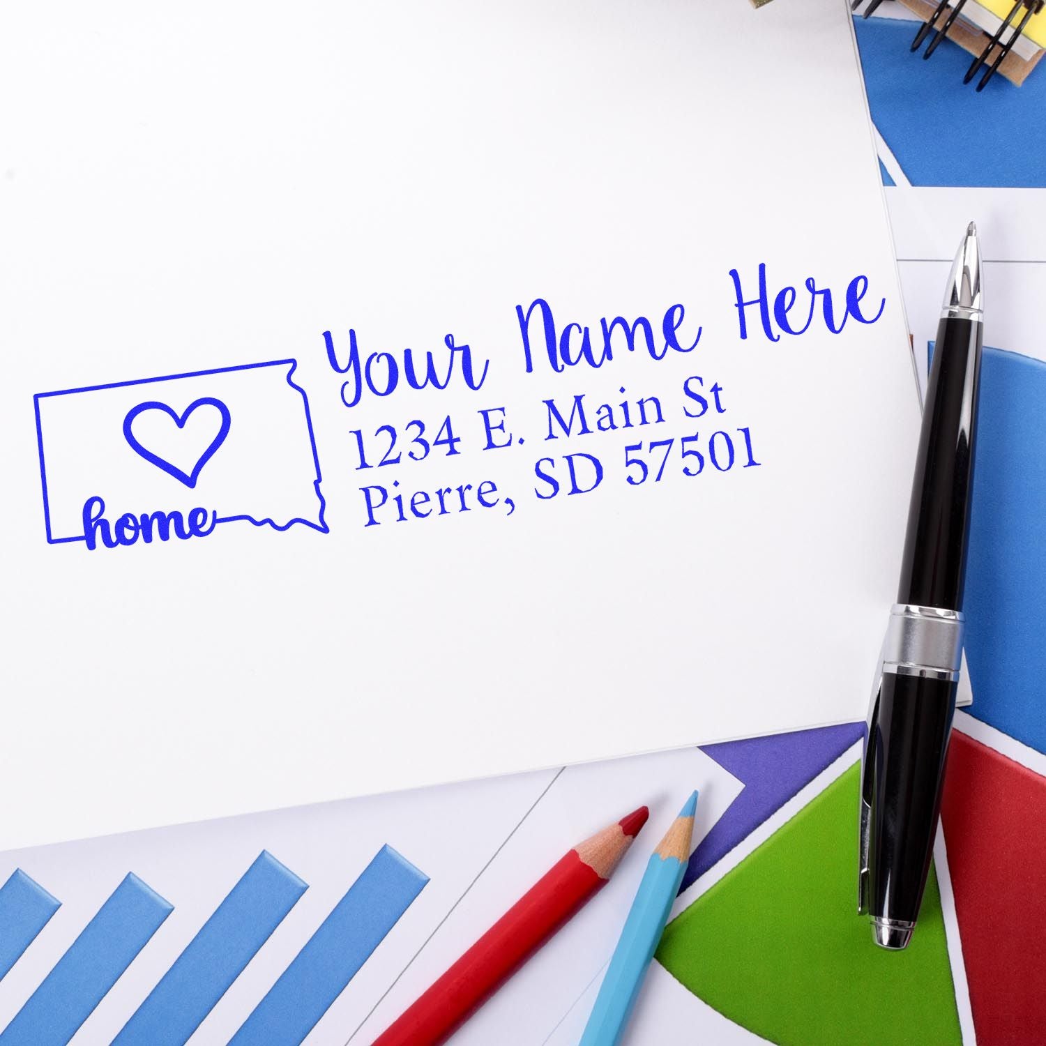 Slim Pre-Inked South Dakota State Love Custom Address Stamp on white paper with blue ink, featuring a heart and home text. Surrounded by colorful papers, a pen, and pencils.