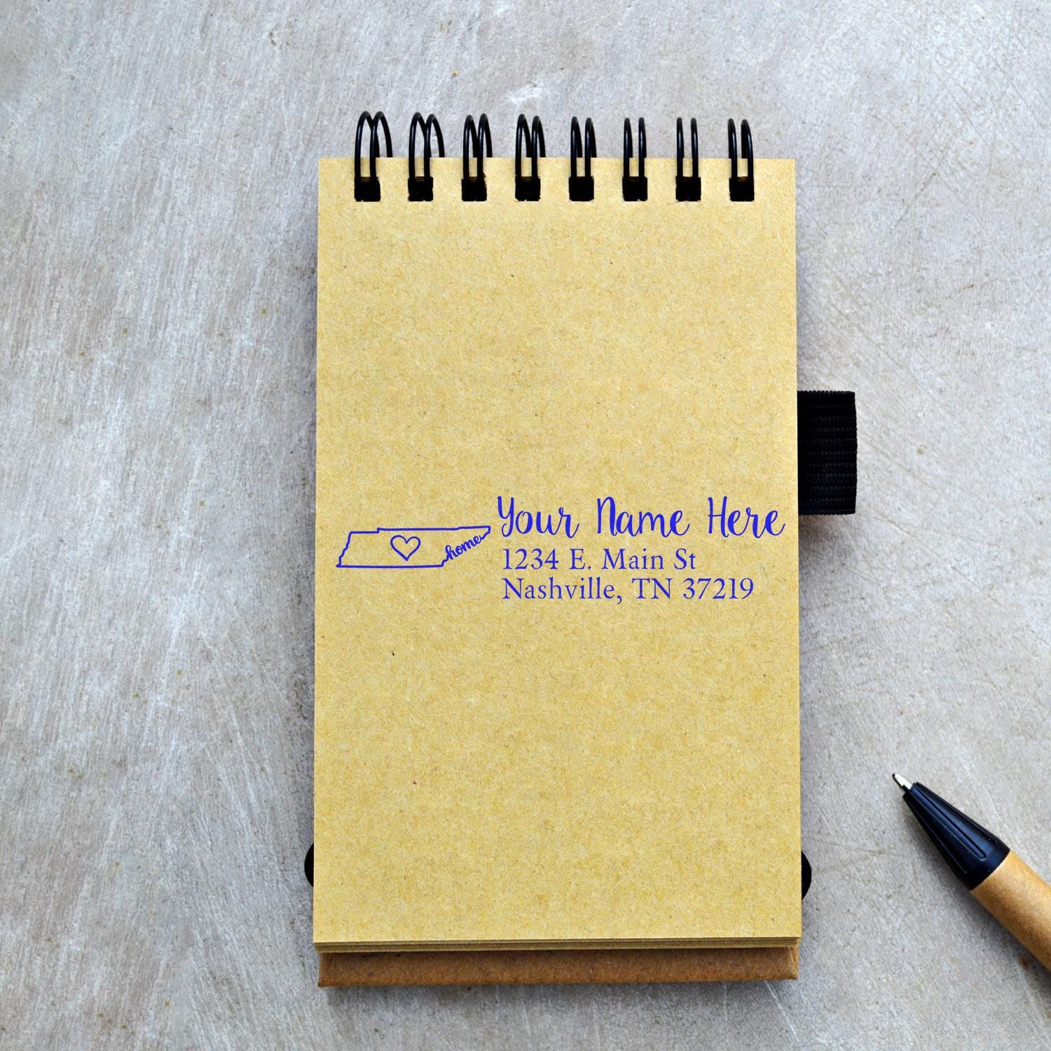 Wood Handle Tennessee Custom Address Rubber Stamp on a notebook, displaying a sample address with a state outline. A pen lies nearby on a textured surface.