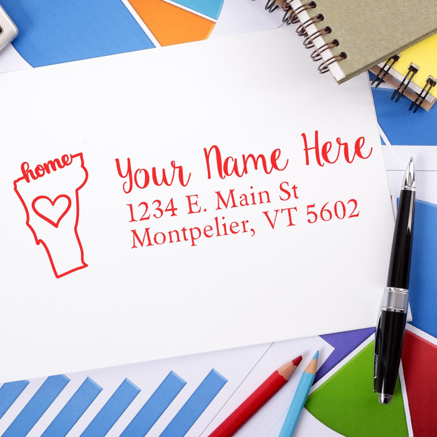 Wood Handle Vermont Custom Address Rubber Stamp on white paper with red ink, surrounded by colorful papers, a notebook, and a pen. The stamp displays a sample address in Montpelier, VT.