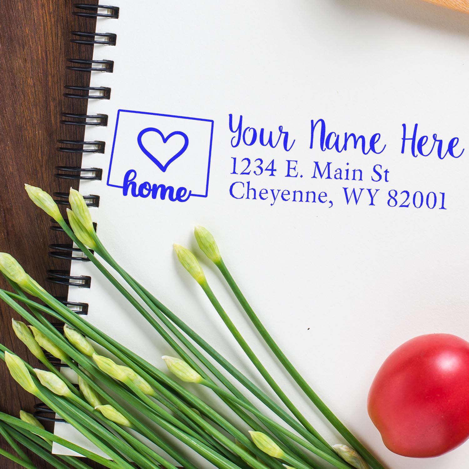 Slim Pre-Inked Wyoming State Love Custom Address Stamp on a notepad with a heart design, surrounded by green stems and a red tomato, showcasing personalized address details in blue ink.