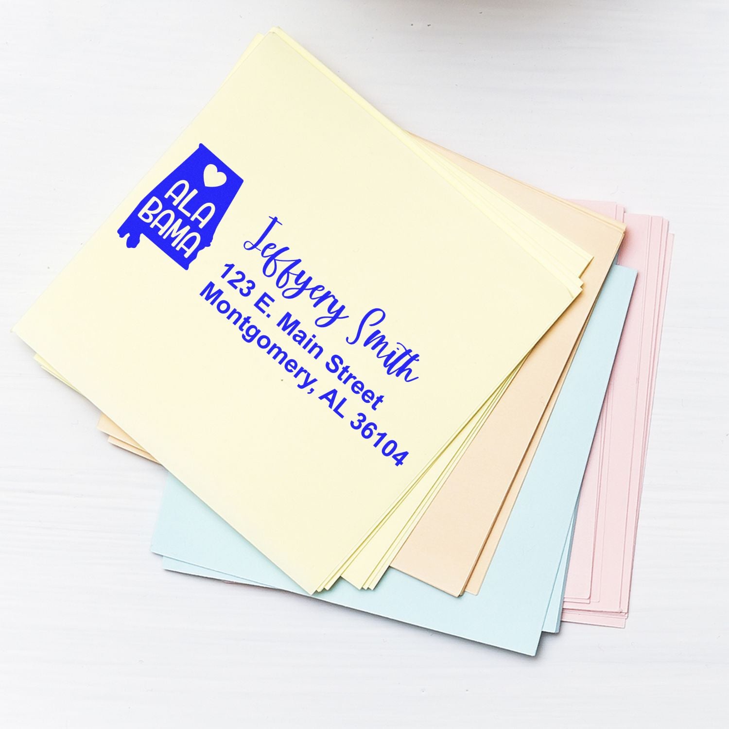 State Love of Alabama Custom Address Stamp Self-Inking used on a stack of pastel envelopes, featuring a blue Alabama state outline with a heart and personalized address in elegant script.