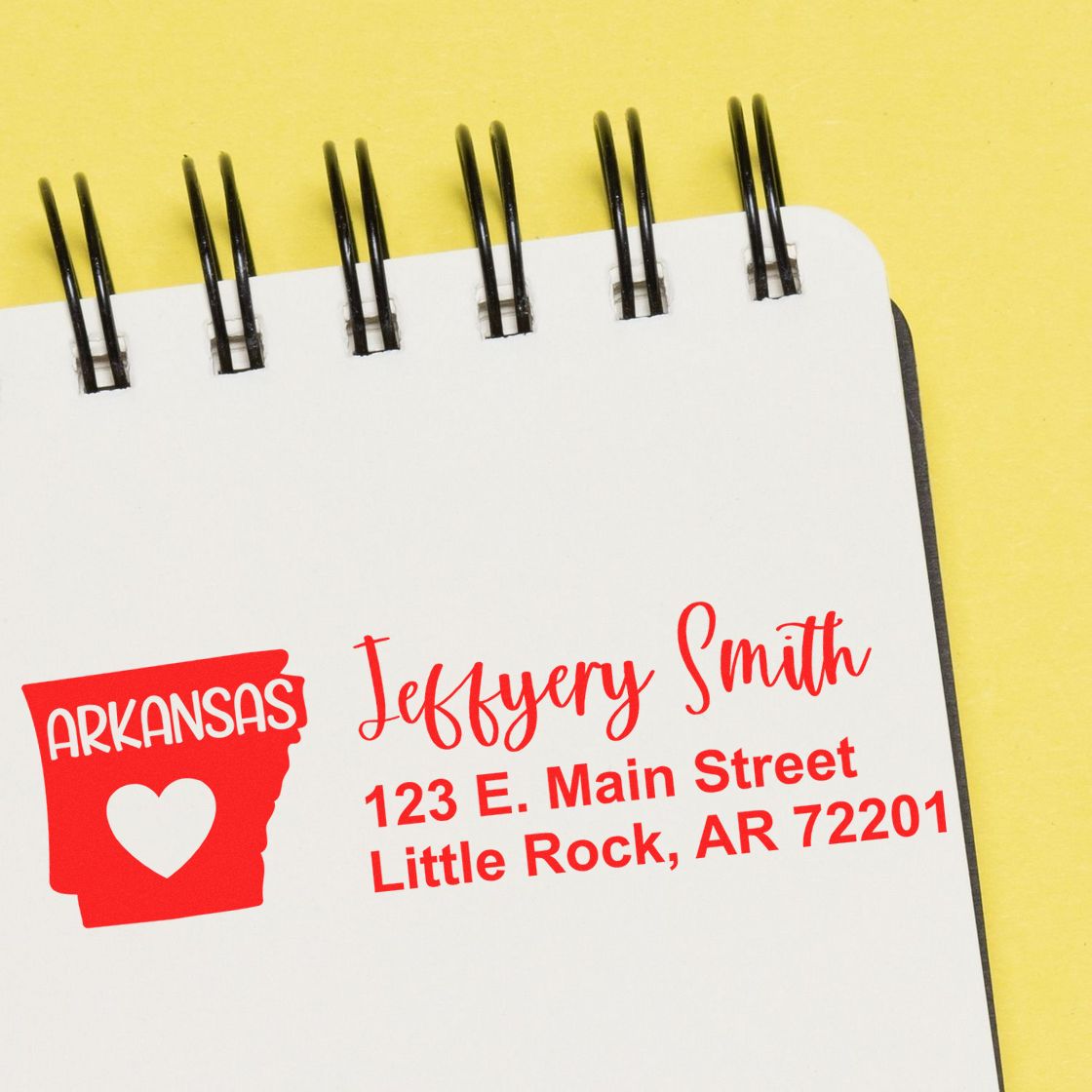 Slim Arkansas Custom Address Stamp for Envelopes displayed on a notepad, featuring a red Arkansas state outline with a heart and personalized address in bold red text.